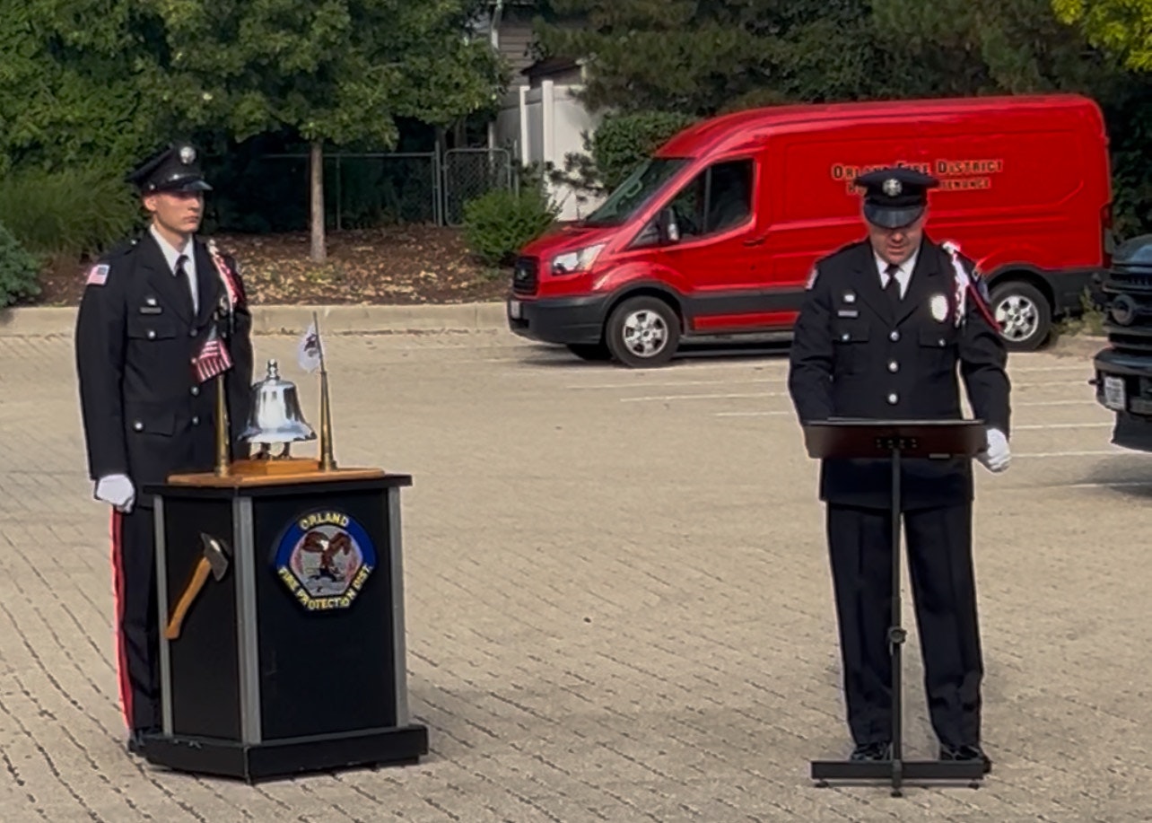 Two uniformed fire officials stand in a parking lot, one by a podium with a bell and flags, the other reading from a lectern.
