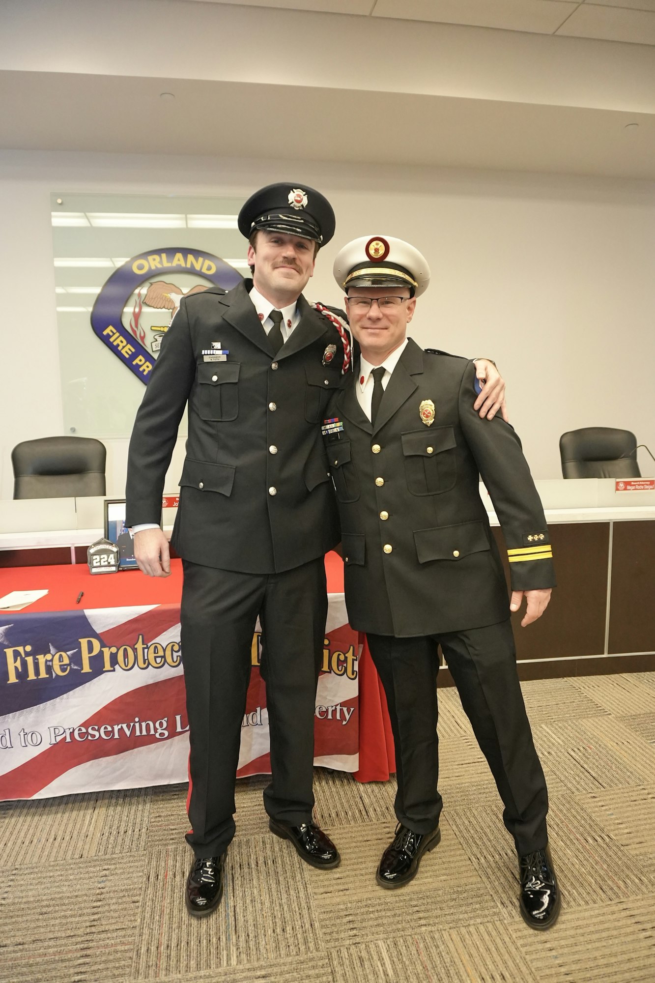 Two firefighters in uniform posing together, smiling in front of a "Fire Protection District" banner.