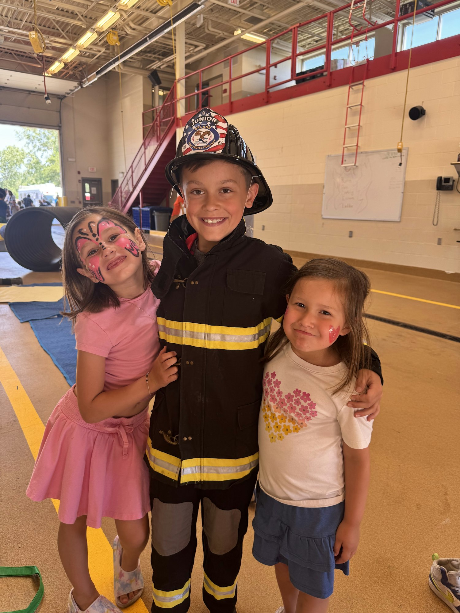 Three kids posing; one in a firefighter costume, another with face paint, and a third smiling. Indoors with gym equipment visible.