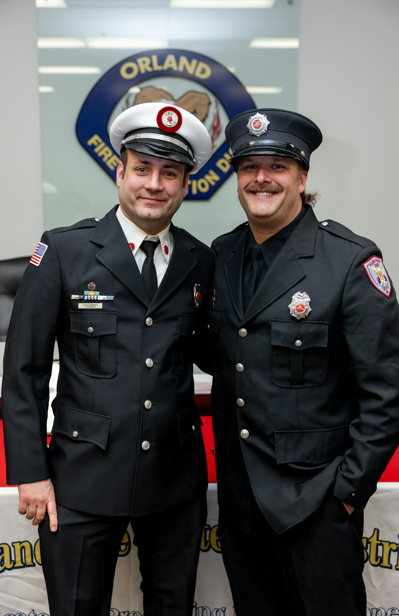 Two firefighters pose together, wearing uniforms in front of an Orland Fire Protection District backdrop.