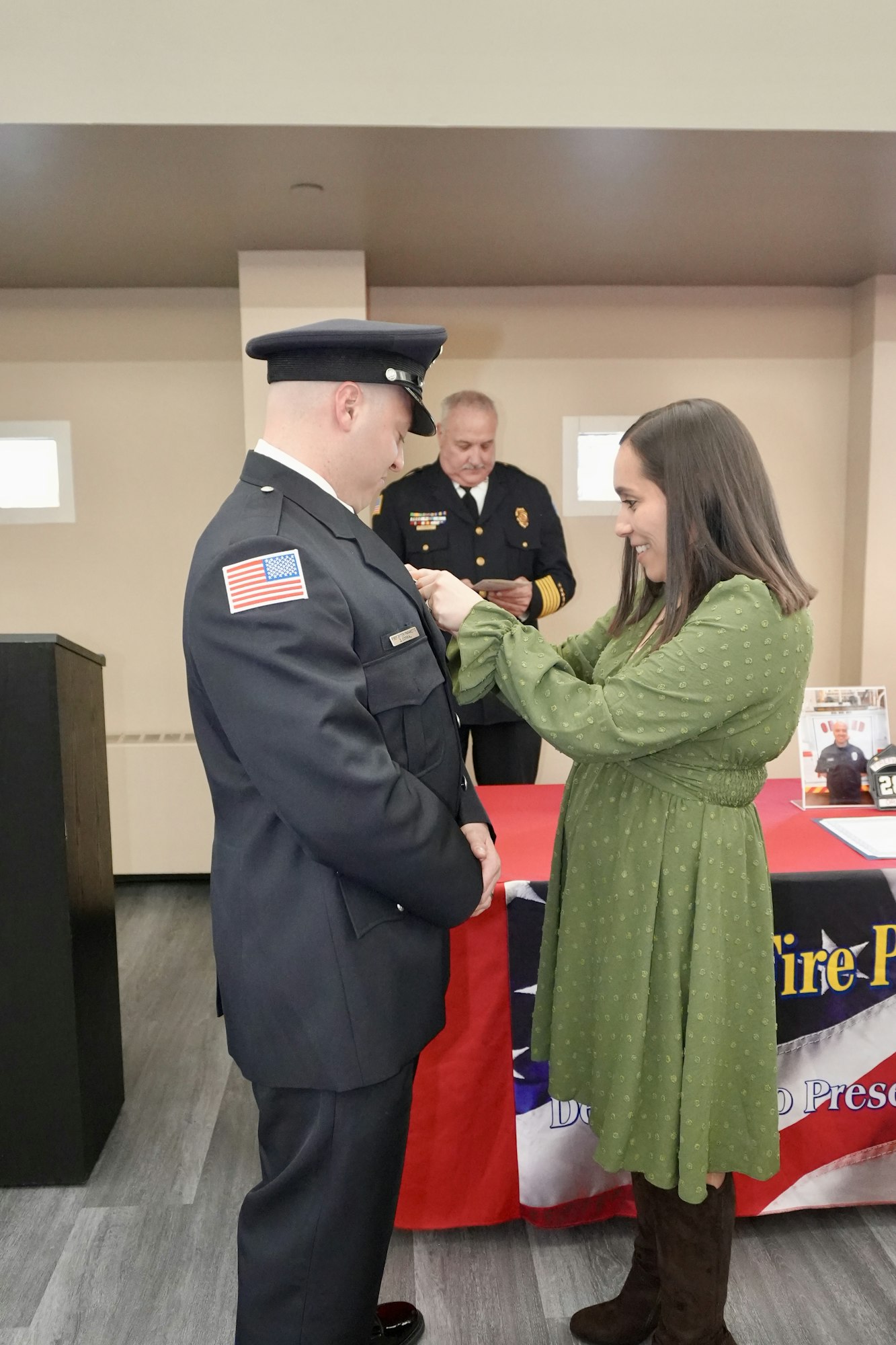 A firefighter in uniform is being pinned by a woman at a ceremony, with another person standing behind them.
