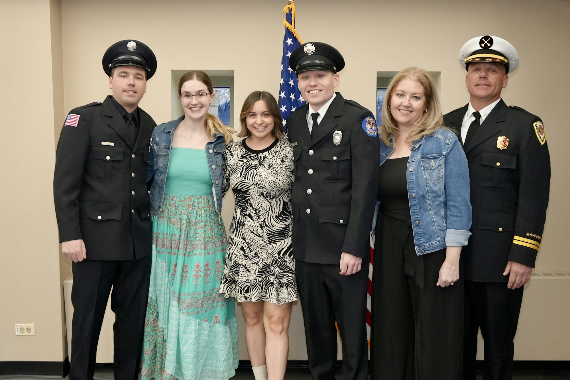 A group of six people, including firefighters in uniform and three women, standing in front of an American flag.