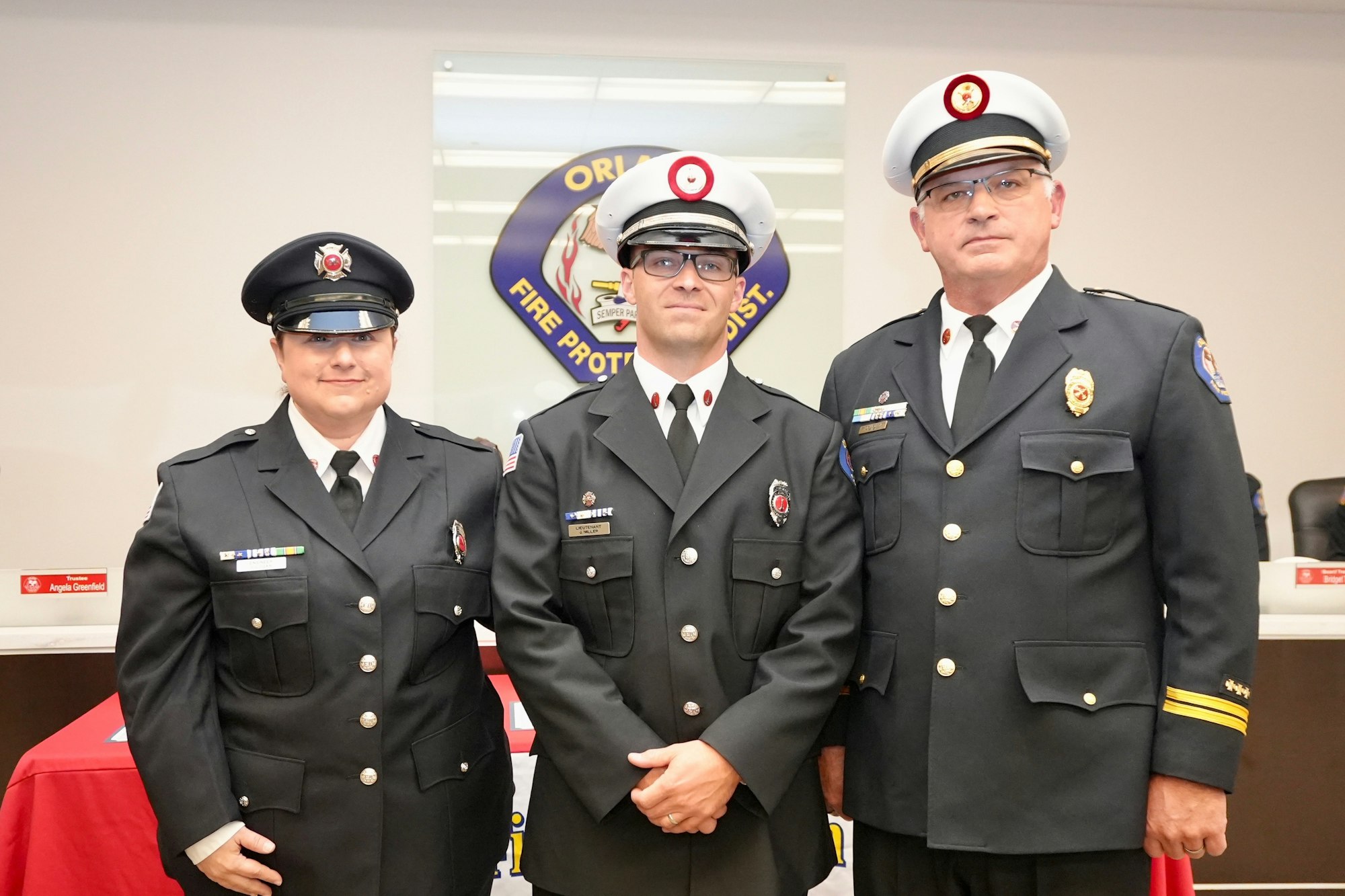 Three people in formal uniforms standing in front of a fire protection district emblem.