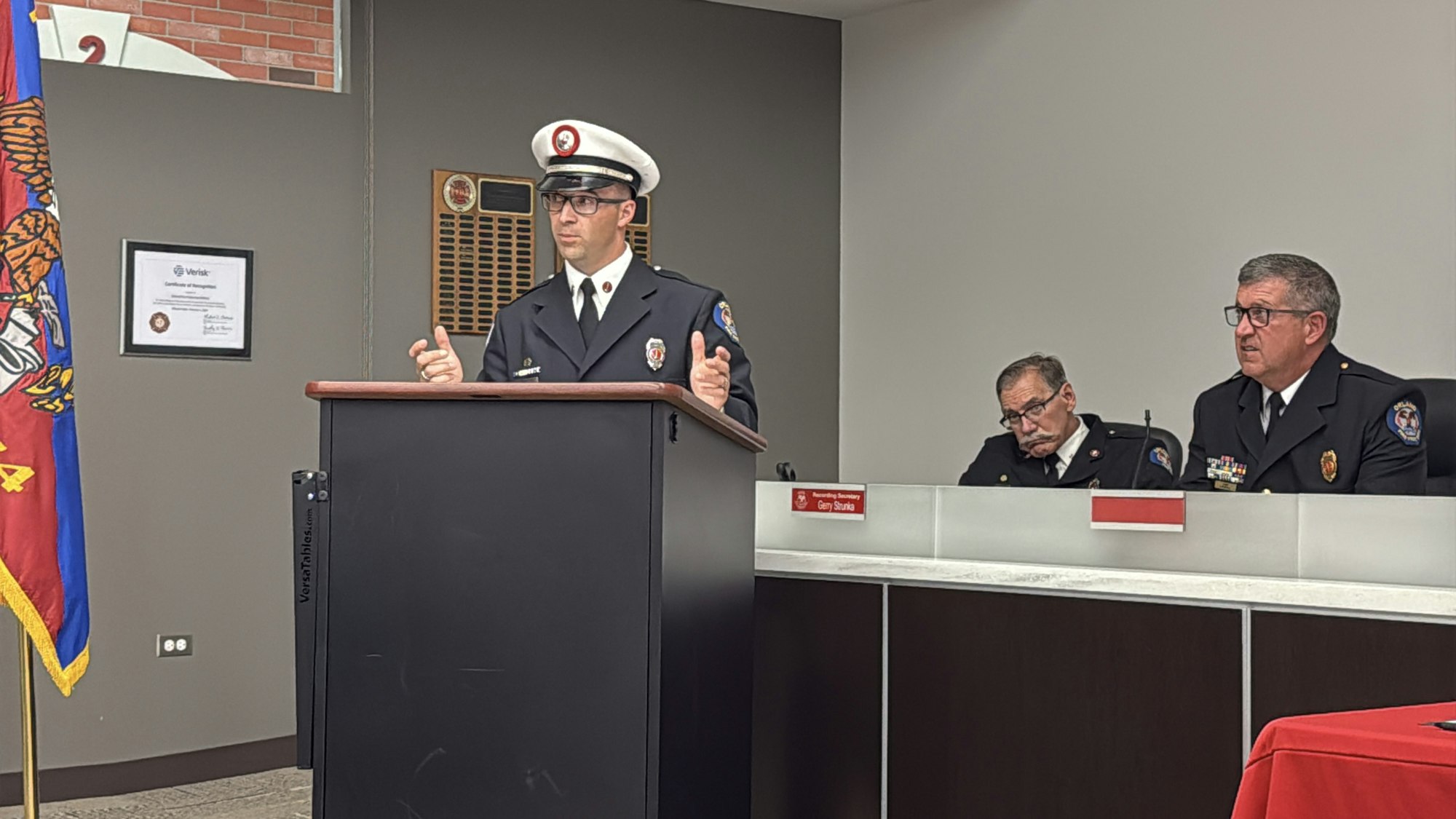 A person in a fire uniform speaks at a podium. Two others sit nearby, also in uniforms, next to a "Recording Secretary" sign.