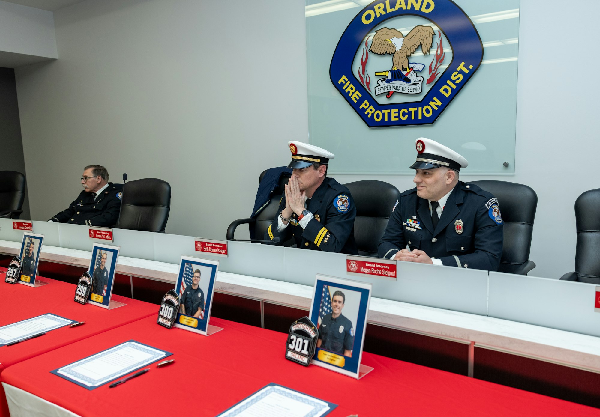 A meeting room with three uniformed fire officials seated at a table, with photos and name tags in front of them.