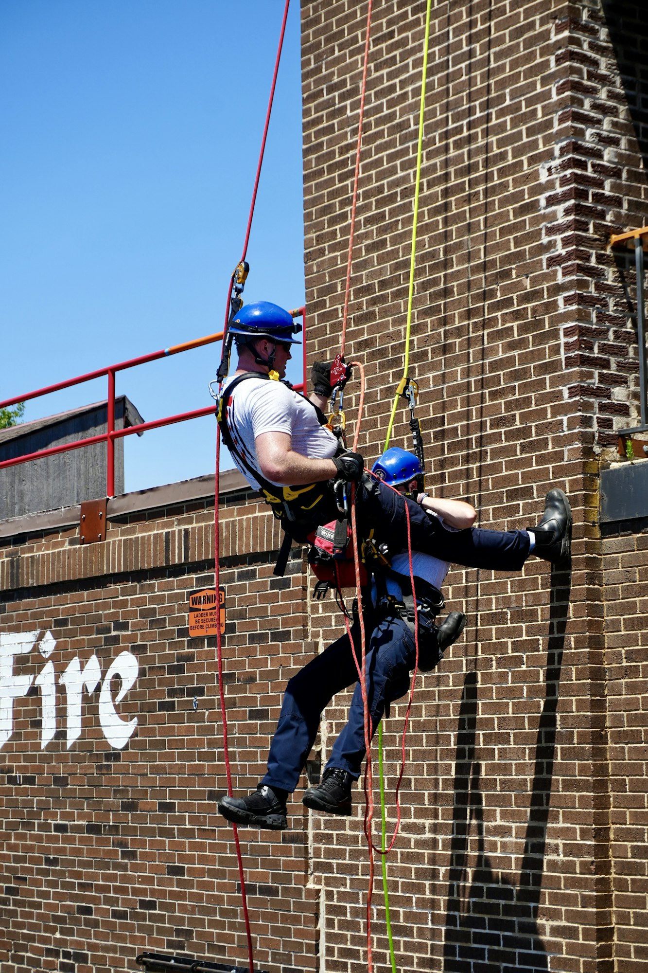 Two people in safety gear performing a rope rescue drill on a brick wall.