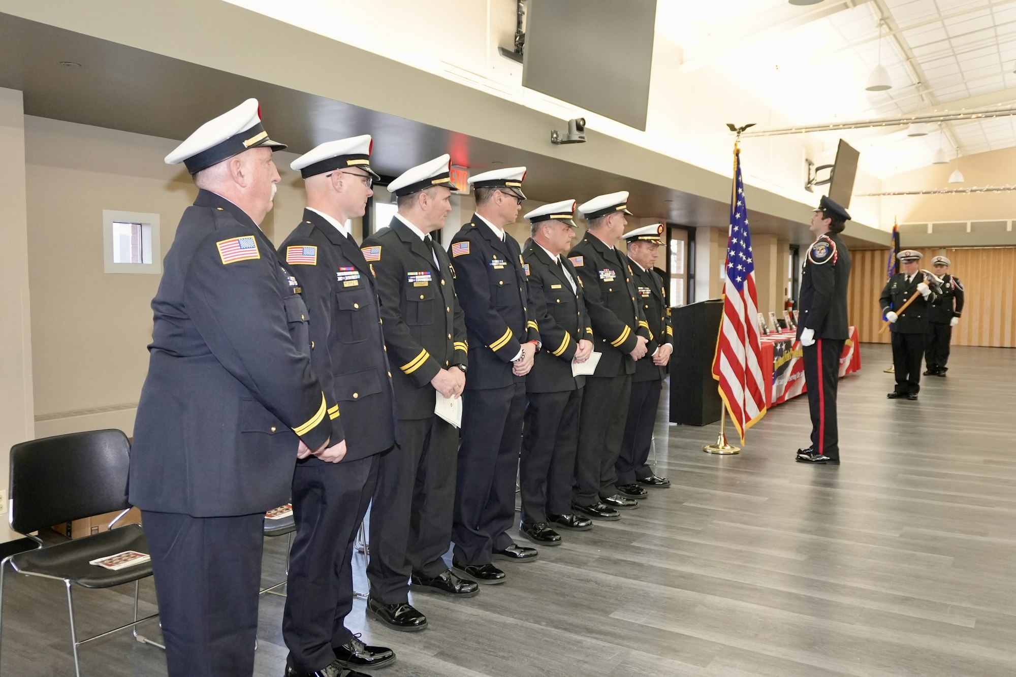 A line of uniformed officers stand facing a podium with the U.S. flag, inside a room with a ceremonial setup.