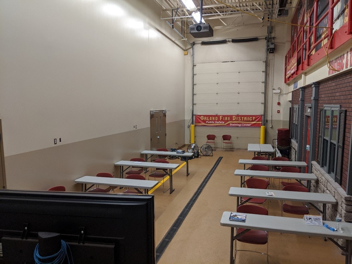 An indoor training facility with tables, chairs, and fire safety equipment; banner reads "Orland Fire District Public Safety Training Center".
