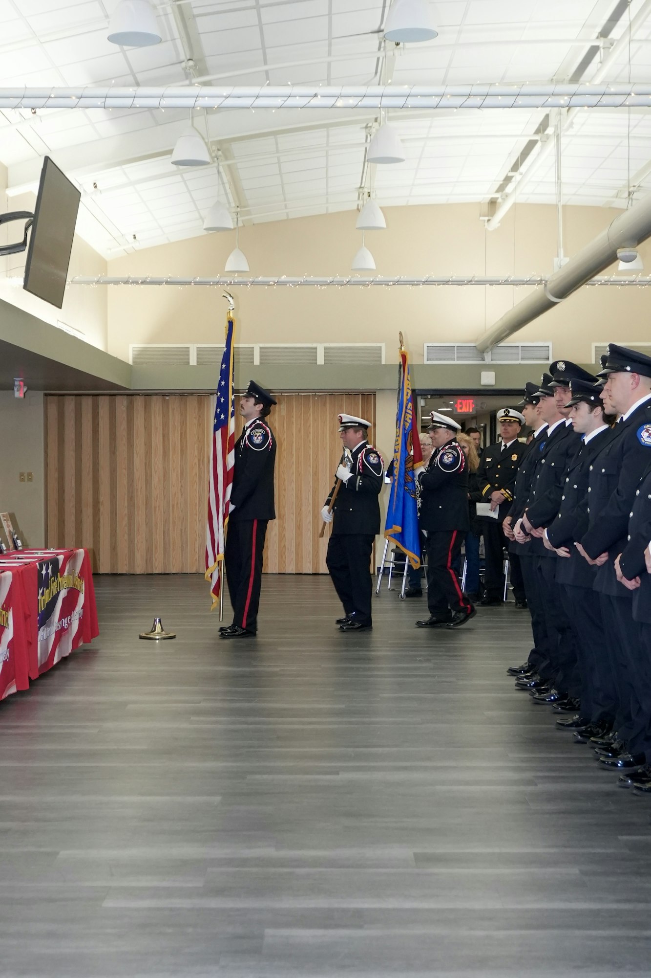 Uniformed personnel in a line, two holding flags, standing inside a room with red-covered tables.