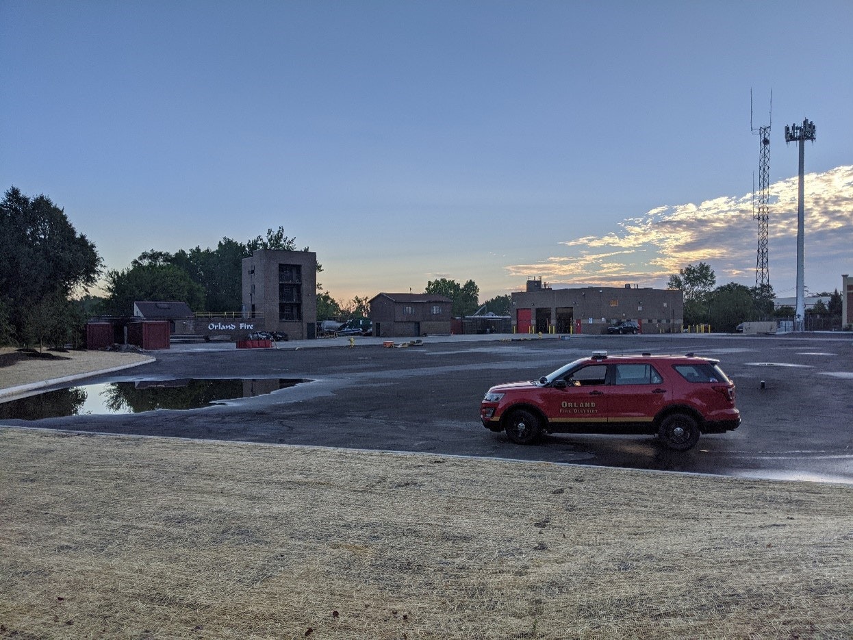 A red emergency vehicle, fire training tower, fire station, antenna tower, trees, and a sunrise or sunset sky.