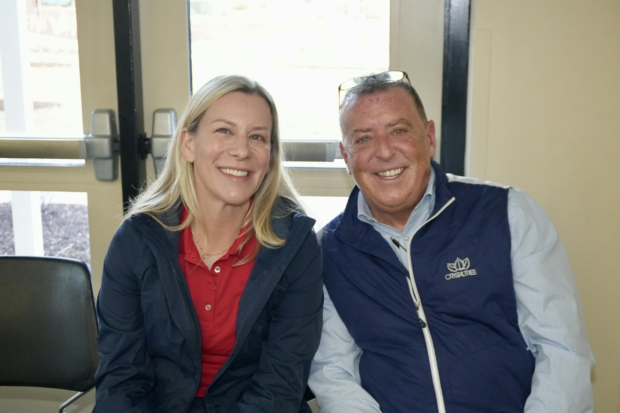 A man and a woman sitting and smiling in front of a window, wearing casual jackets.