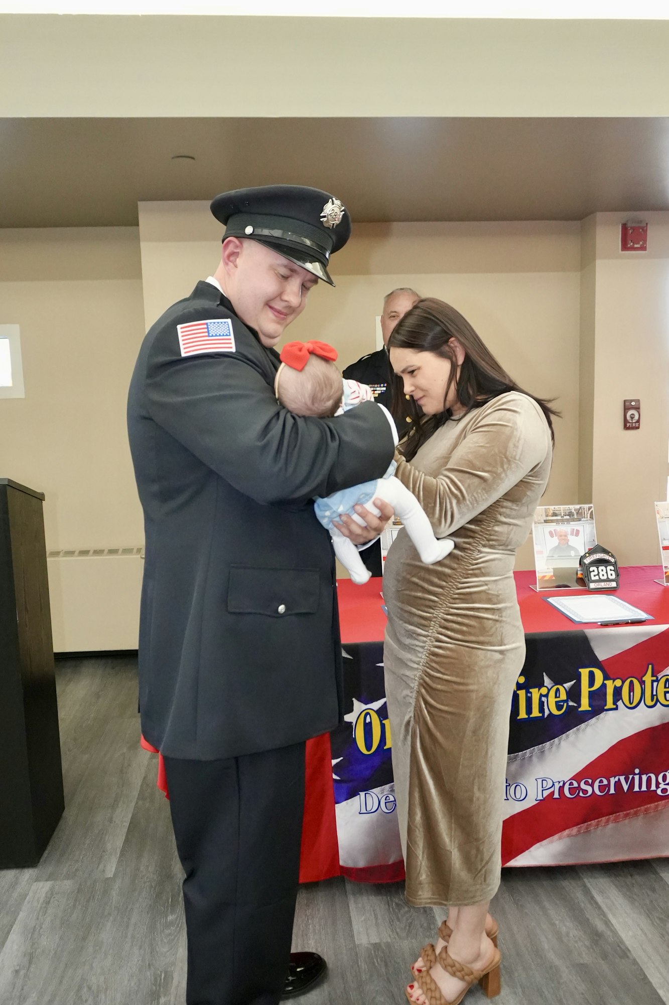A firefighter in uniform holds a baby with a red bow as a woman looks on. There's a table with a patriotic-themed cloth in the background.