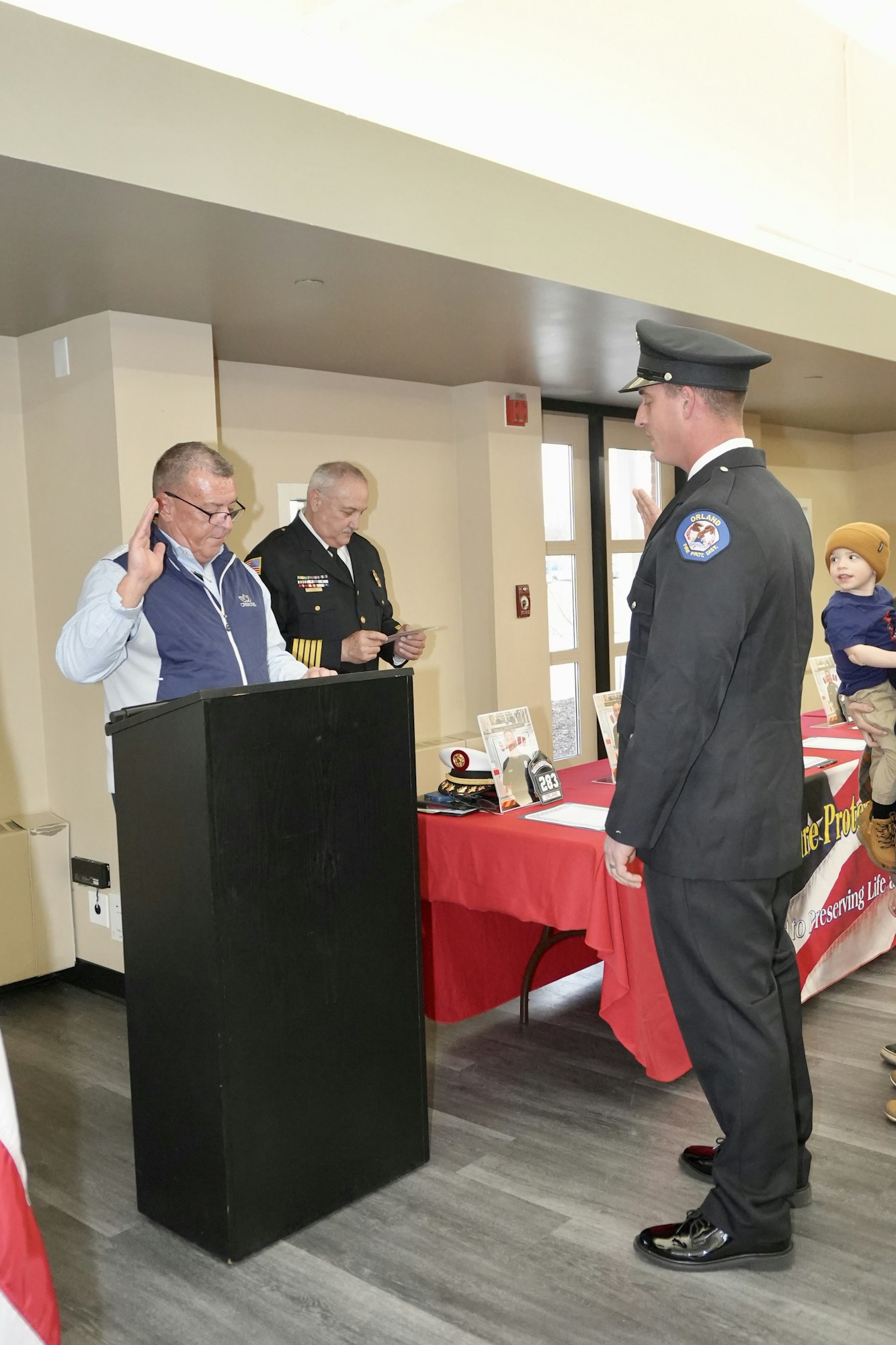 A firefighter is being sworn in at a ceremony with officials at a podium, while another person and a child look on.