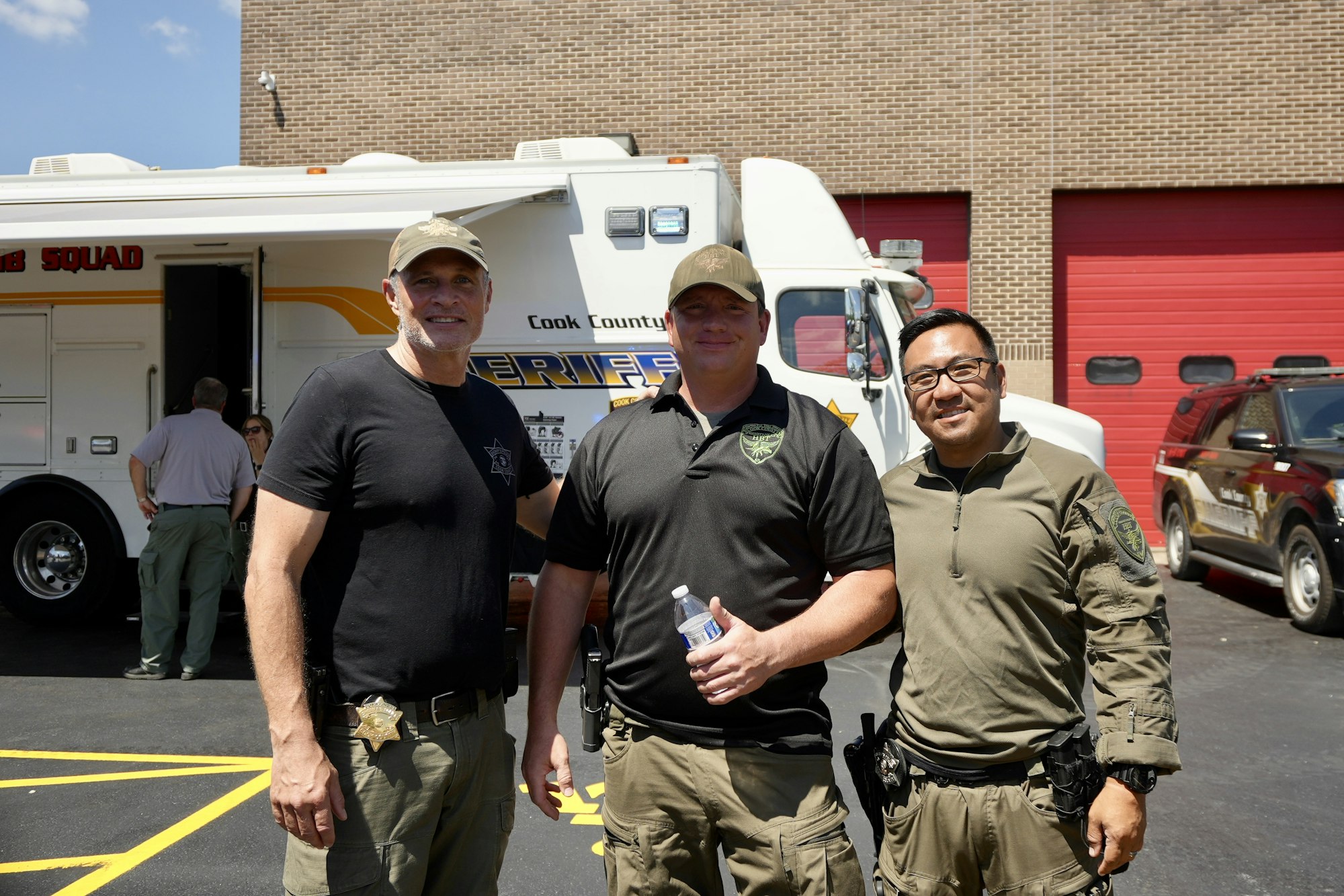 Three uniformed men standing in front of a sheriff's vehicle, smiling.