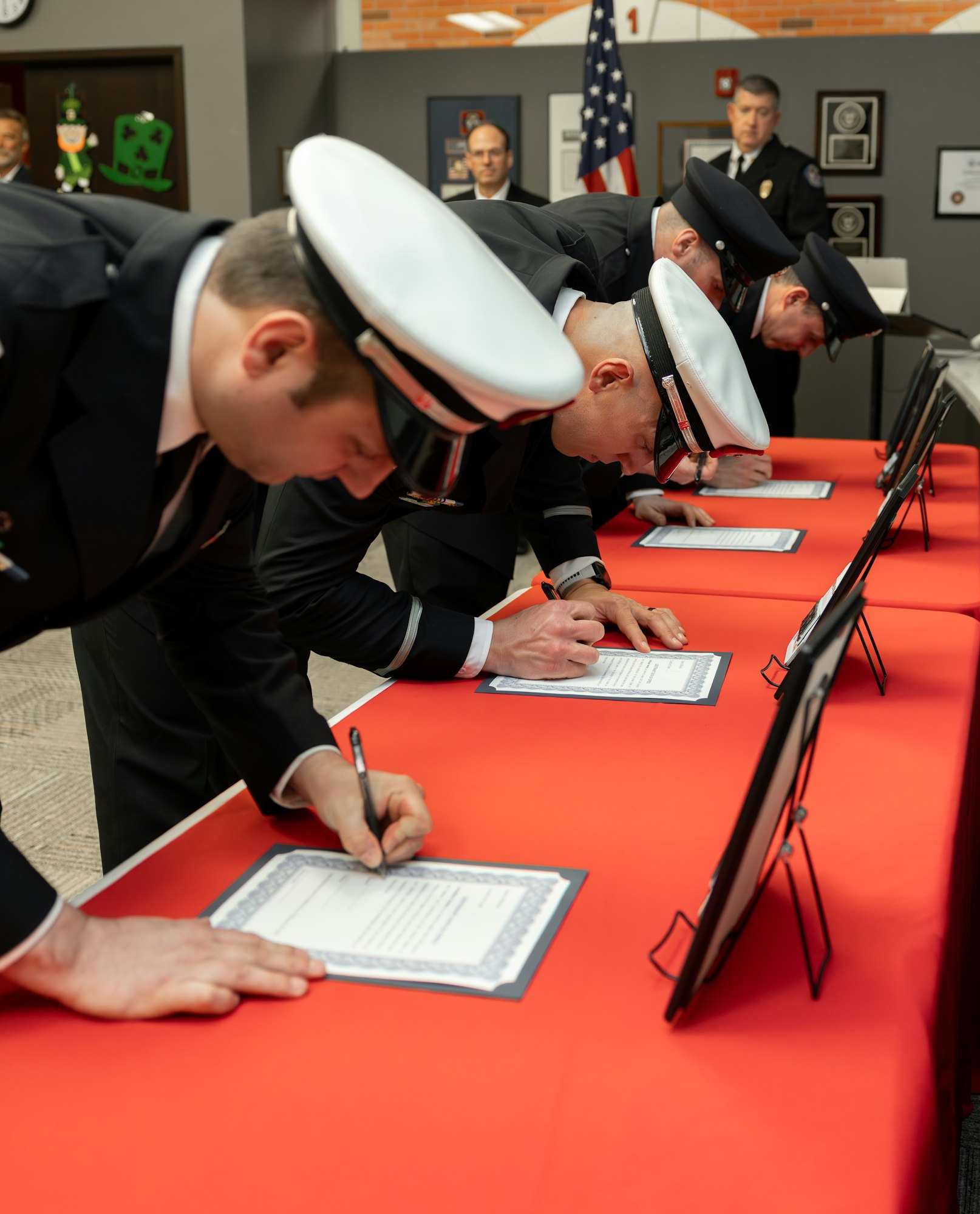 Uniformed individuals are signing documents on a red table, with an American flag and other attendees in the background.