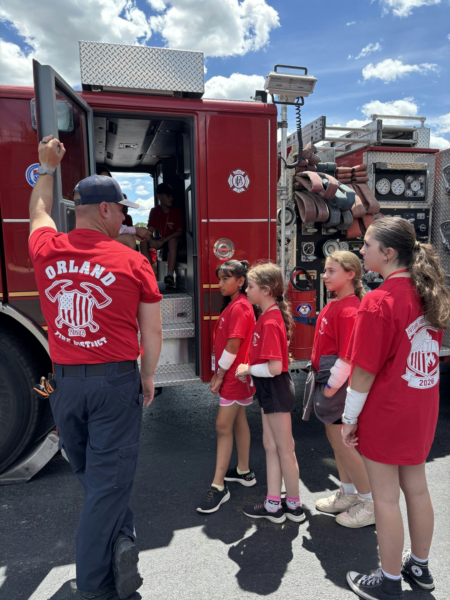 A group of children in red shirts tours a fire truck, guided by a firefighter on a sunny day.