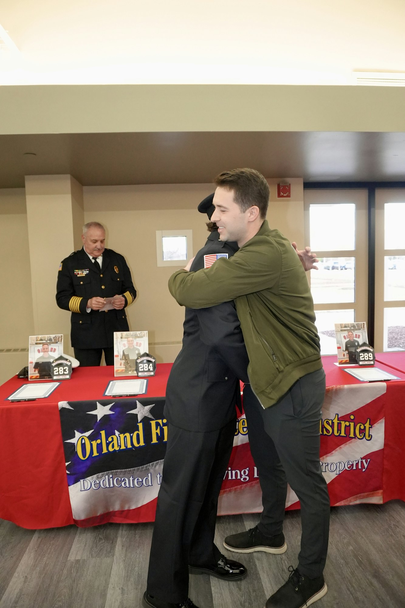Two people hugging in front of a table with firefighter gear; a man in uniform stands behind.