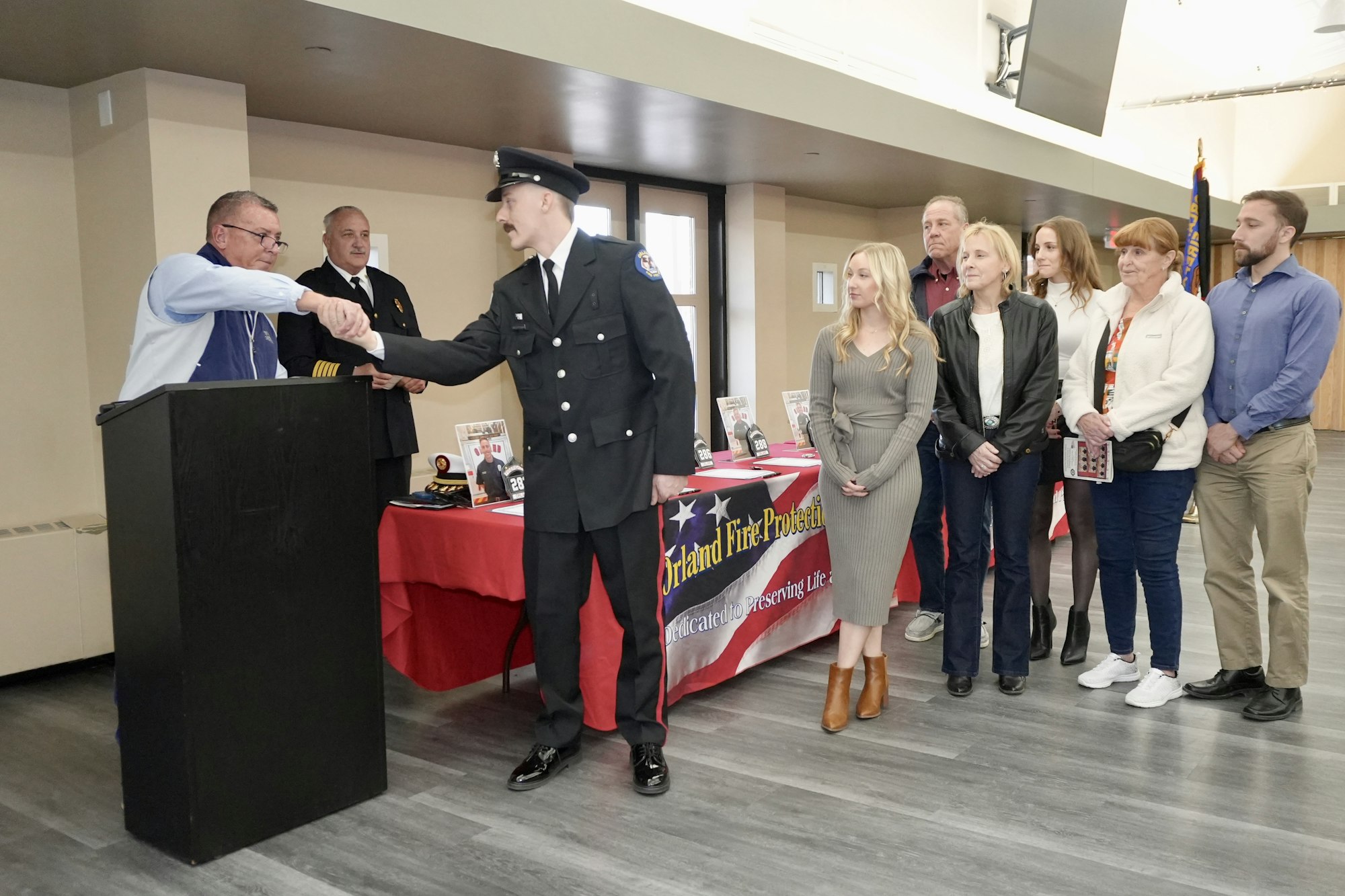 A man in a firefighter uniform shakes hands at a podium while people stand nearby. A table has an "Orland Fire Protection" banner.
