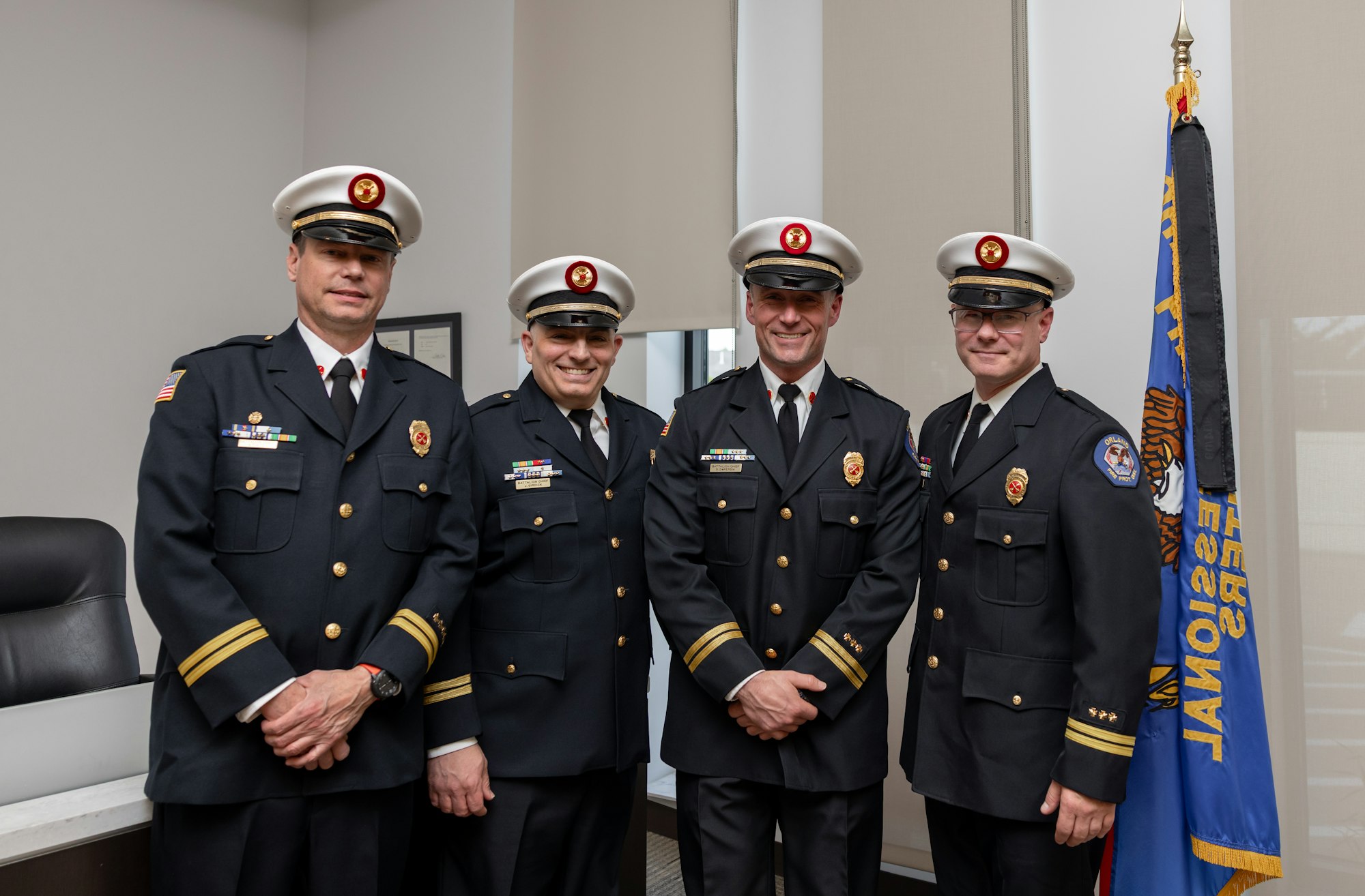 A group of four uniformed officers posing together, all wearing hats and badges, with a flag in the background.