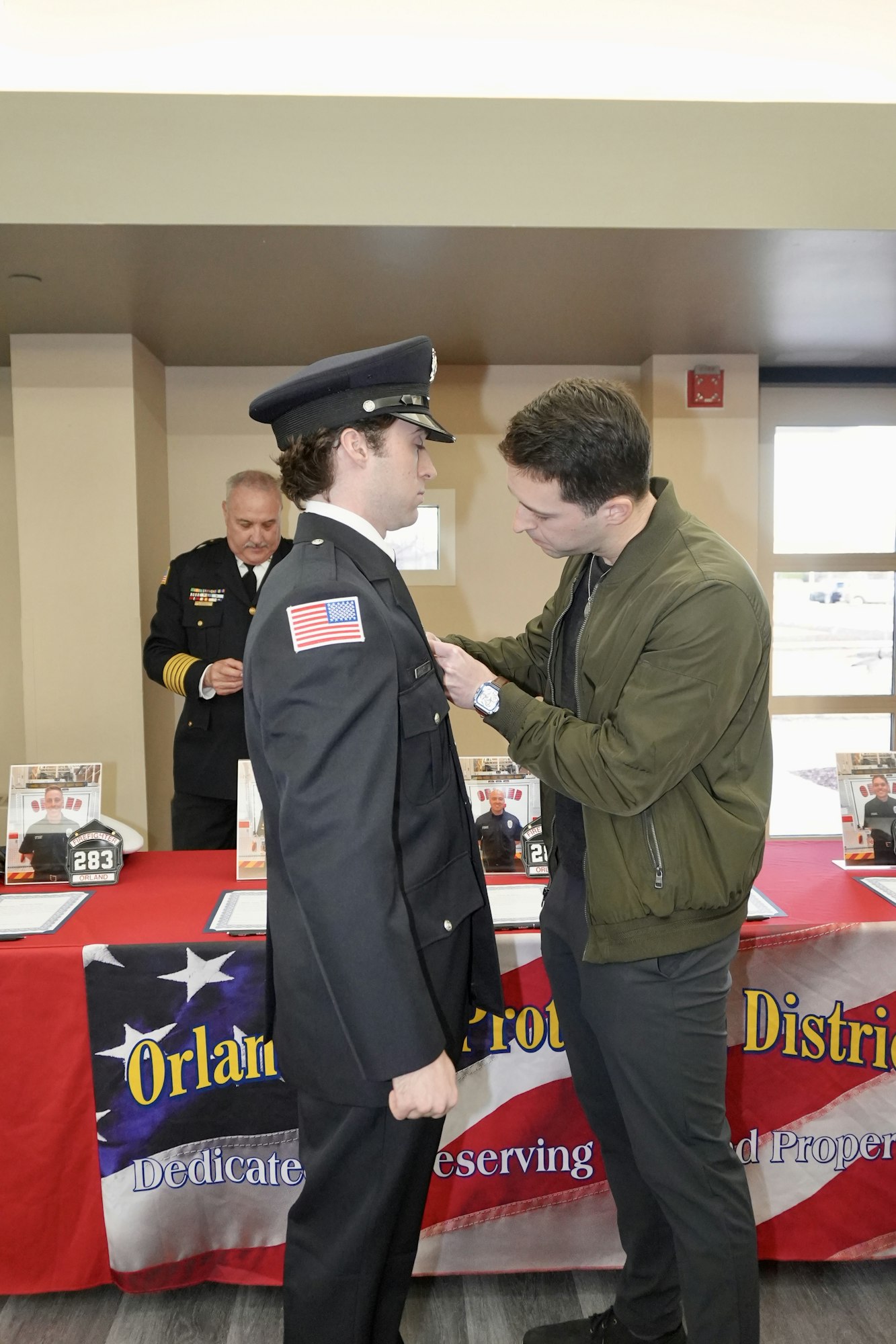 A man pins a badge on a uniformed officer's chest, with another uniformed man in the background at a ceremony or event.