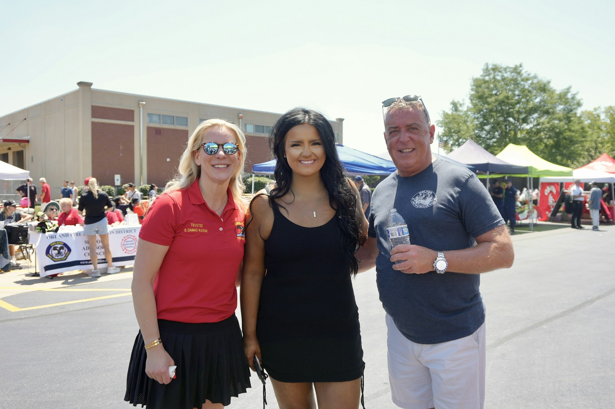 Three people smiling at an outdoor event with tents and a building in the background.