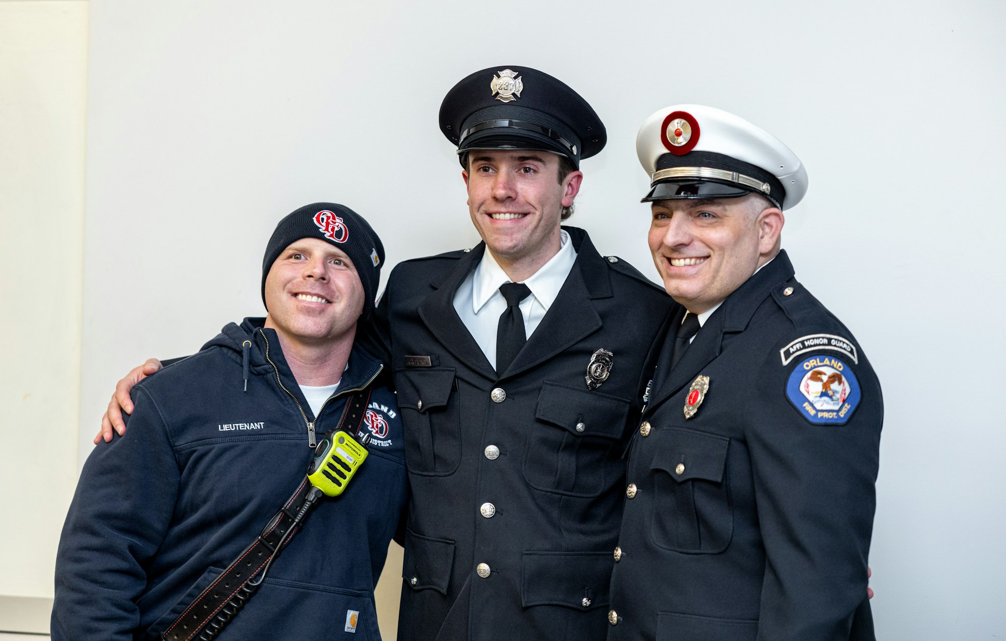 Three men in uniform smile for a photo; one is in a firefighter hoodie, the others are in dress uniforms.