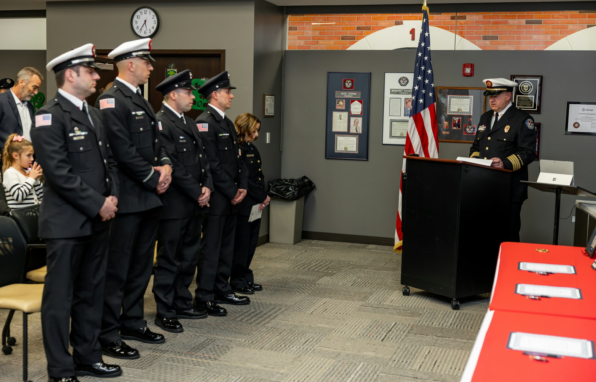 A formal event with uniformed personnel being addressed by a leader, with the American flag and framed honors in the background.