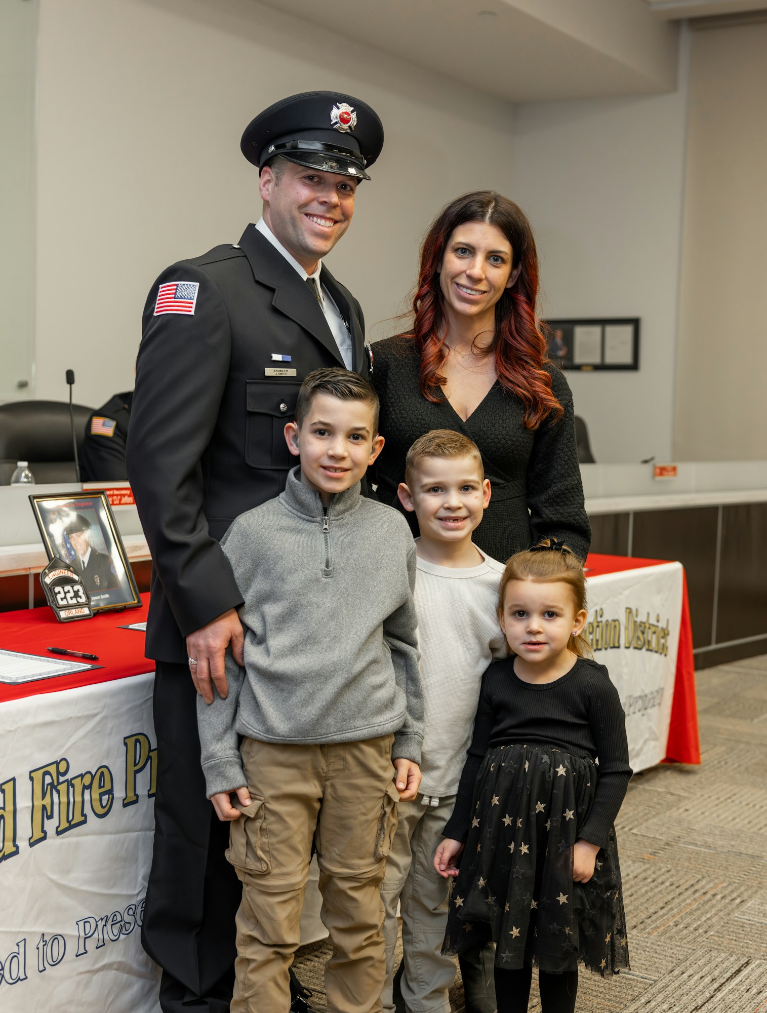 A firefighter in uniform poses with his family, including three children, at an event with a table displaying a fire department logo.