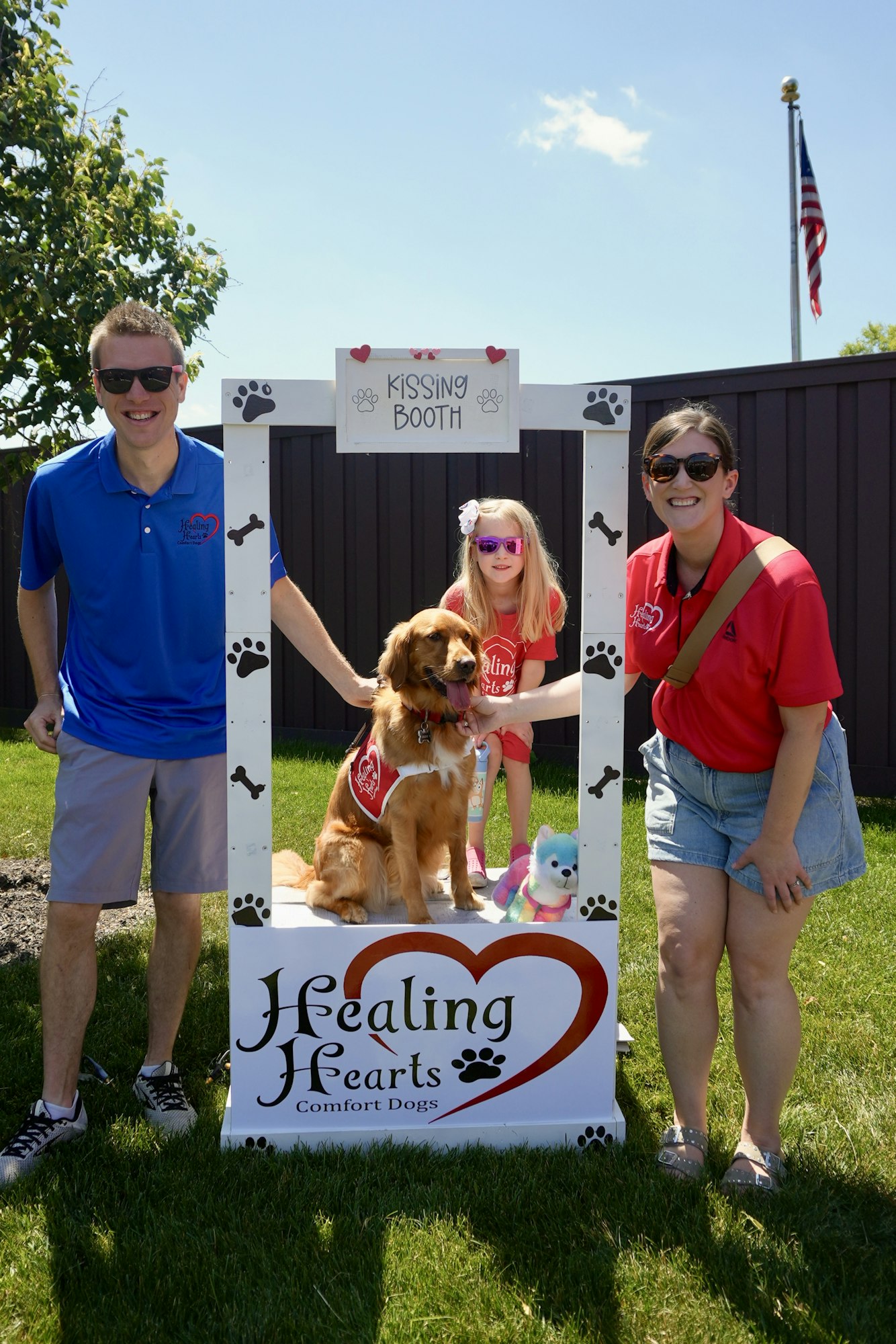 Three people and a comfort dog at a "Kissing Booth" with a "Healing Hearts" sign outdoors.