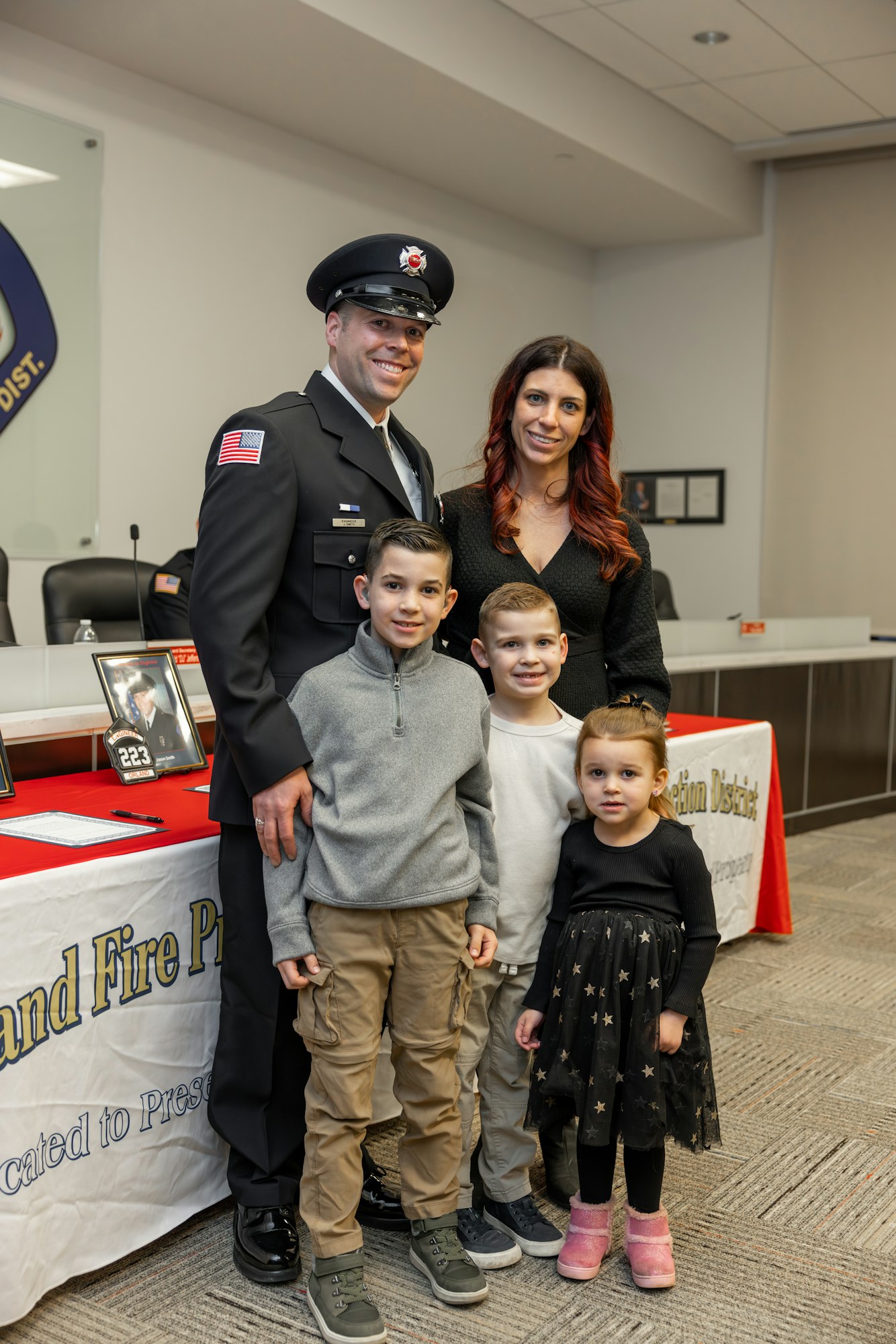 A smiling firefighter stands with his family, including three children, at a ceremony with a display table in the background.
