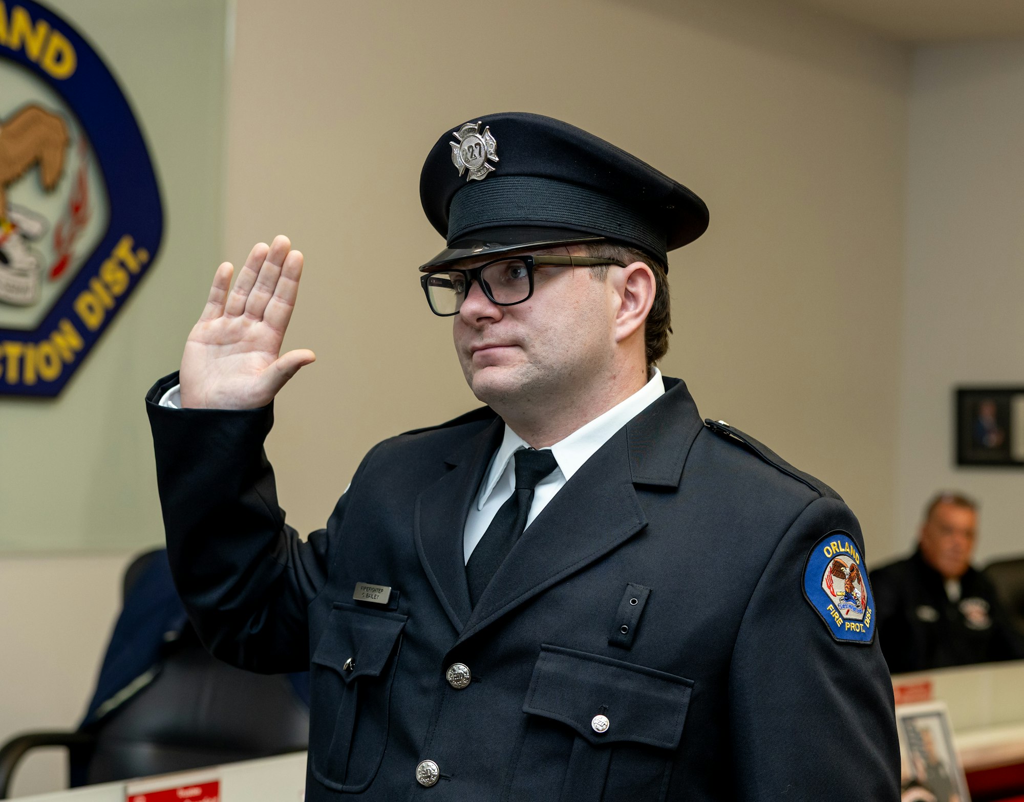 A person in a uniform is taking an oath, likely as part of a ceremony, with others present in the background.