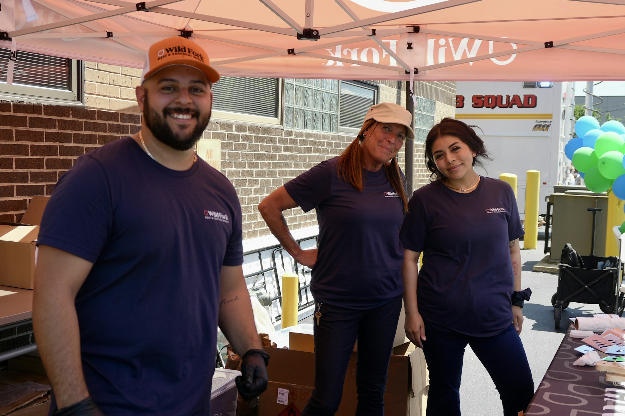 Three people in matching shirts under a tent, smiling at a booth.