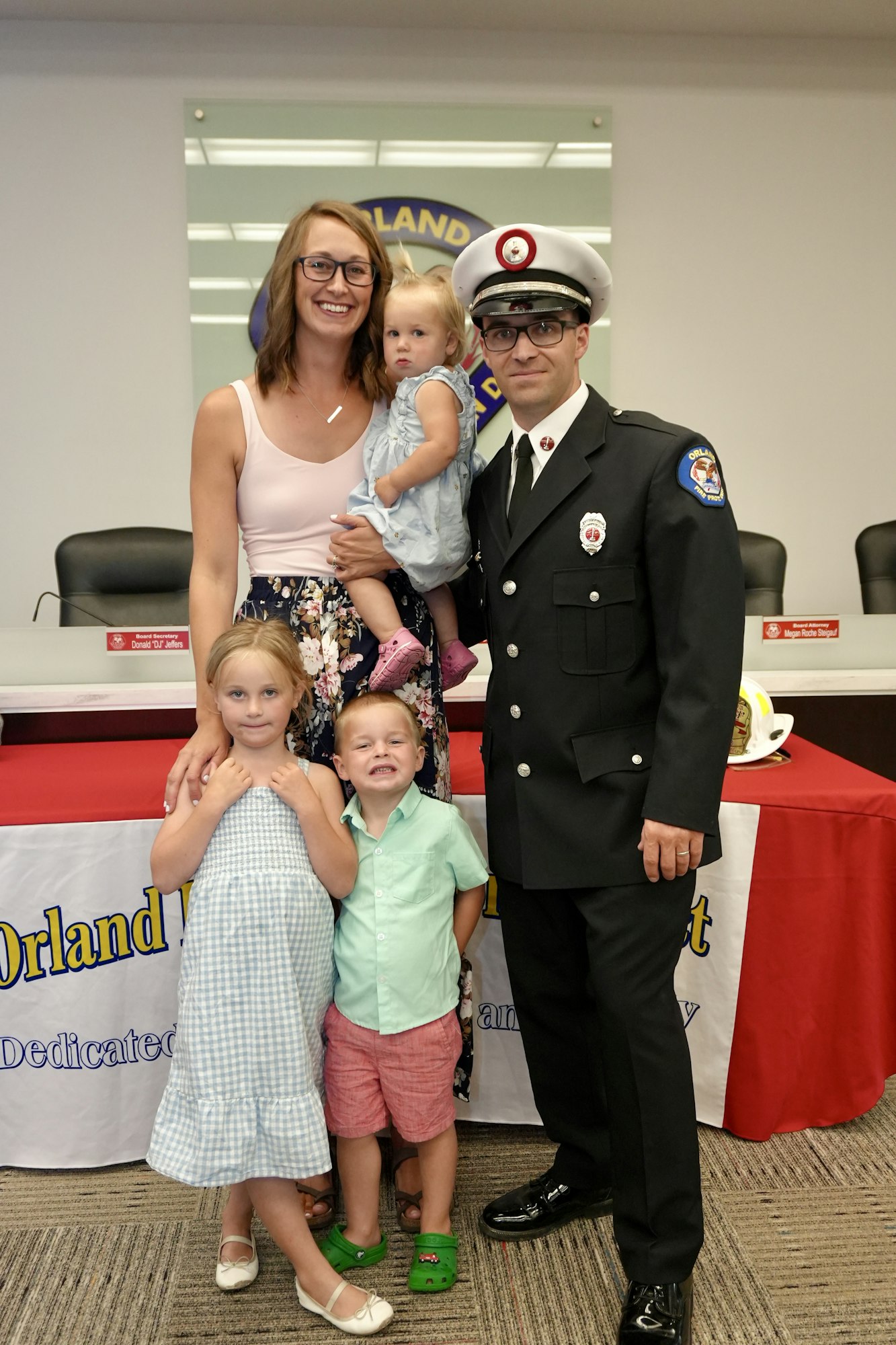 A firefighter in uniform poses with a woman and three children in front of a banner with "Orland" visible.