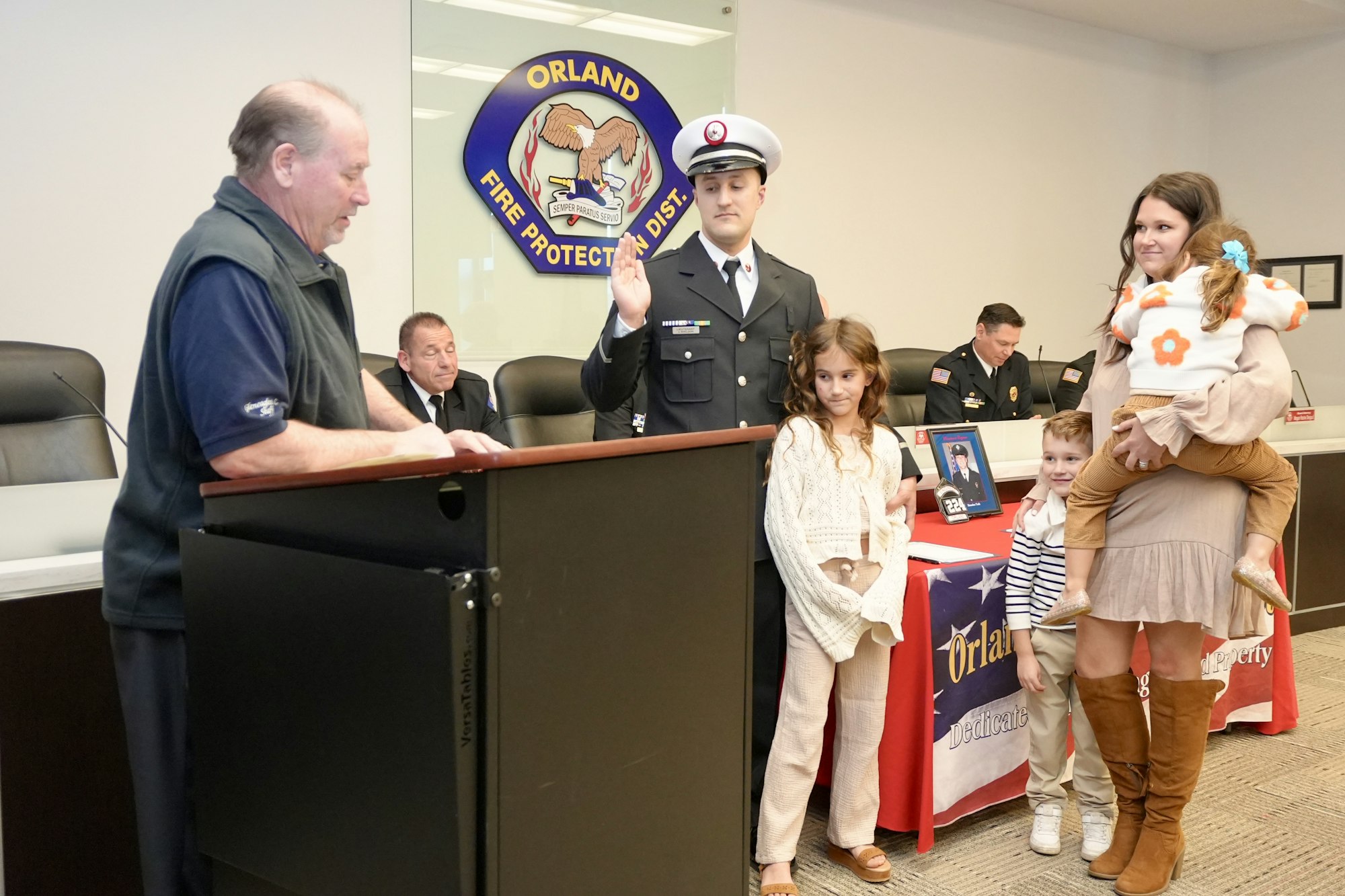 A firefighter is being sworn in at a podium with family and officials present. The Orland Fire Protection District sign is in the background.