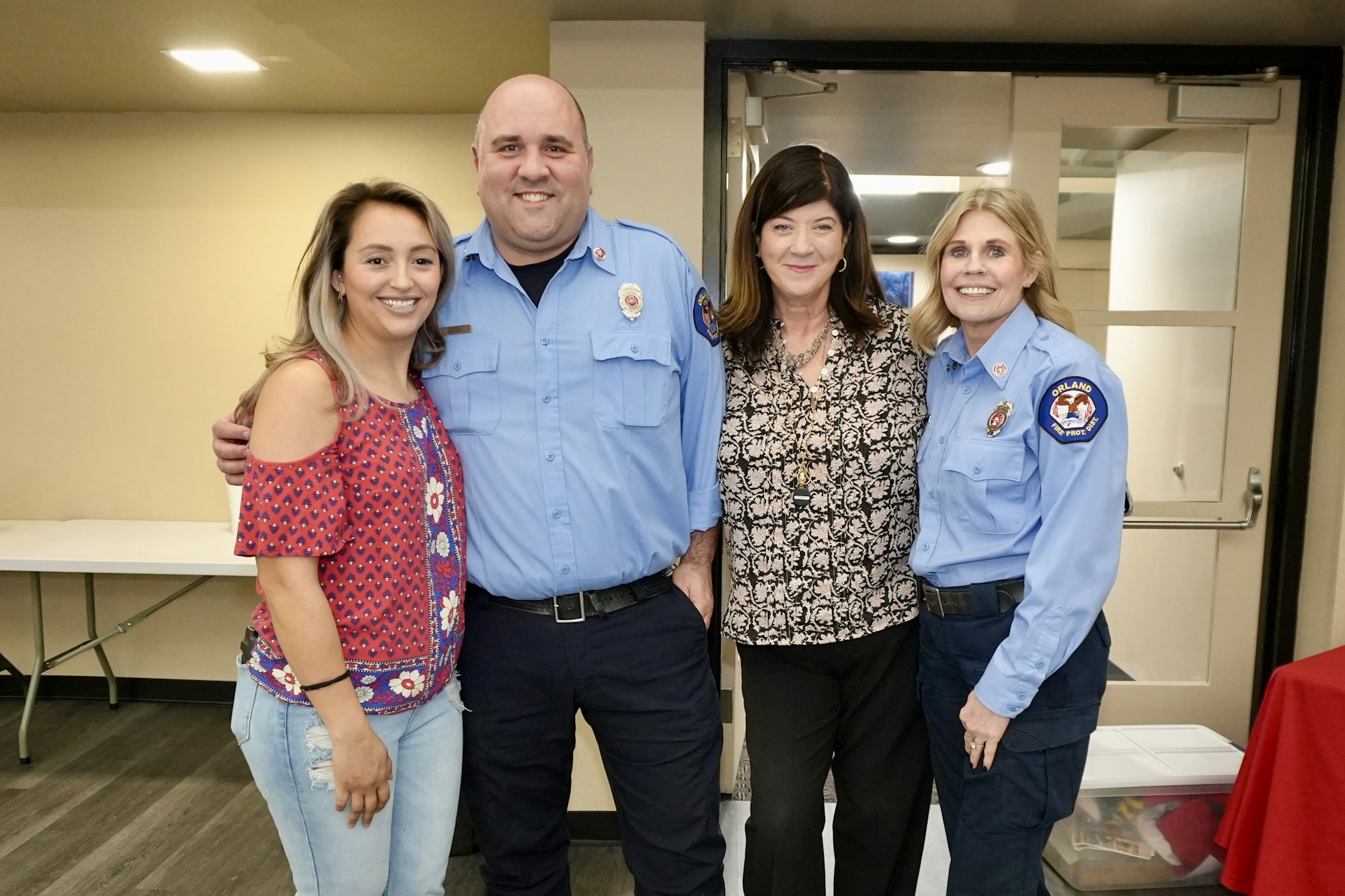 Four people smiling, two in fire department uniforms, standing indoors.