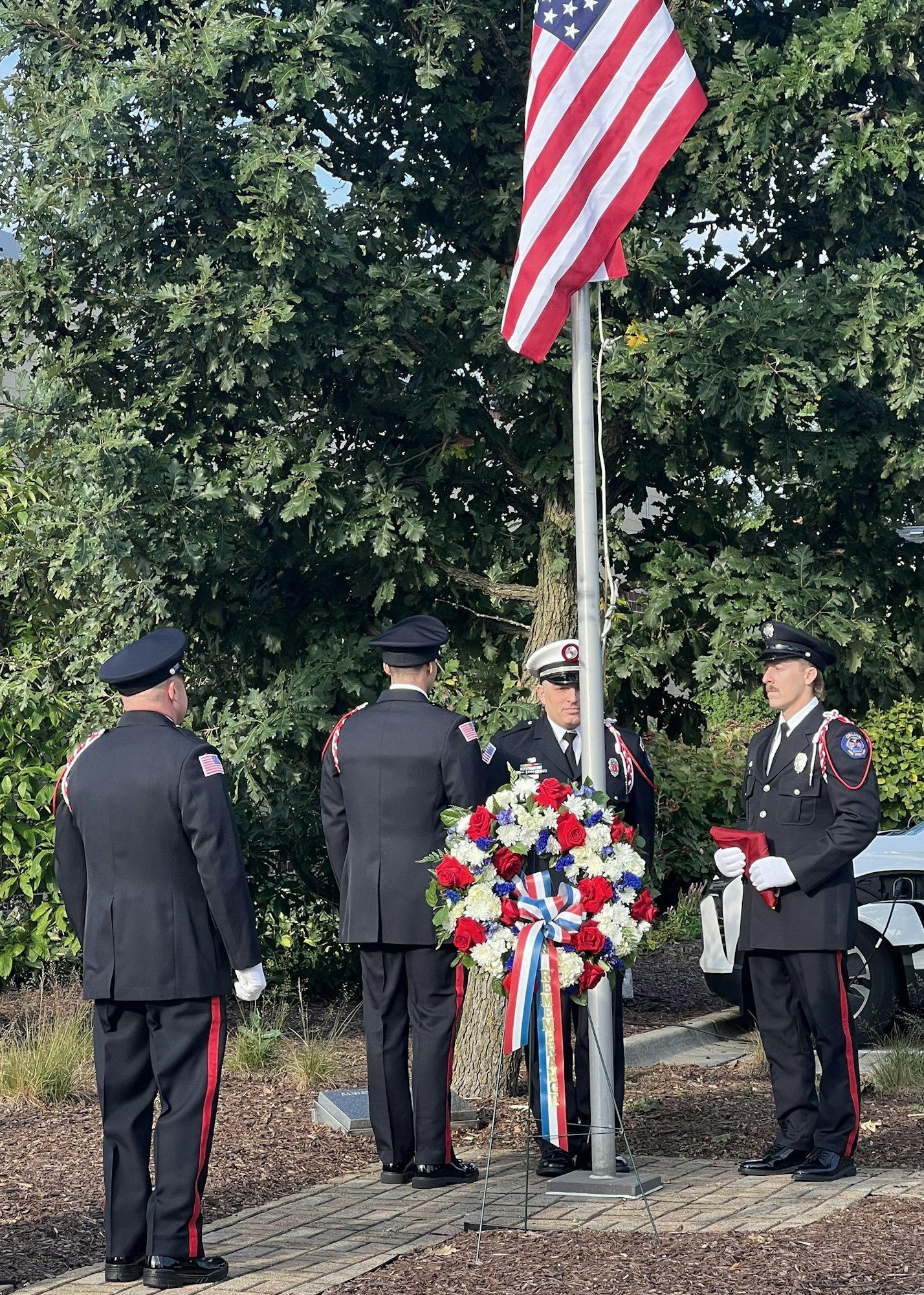 Four uniformed individuals stand around a flagpole with a U.S. flag and a floral wreath.