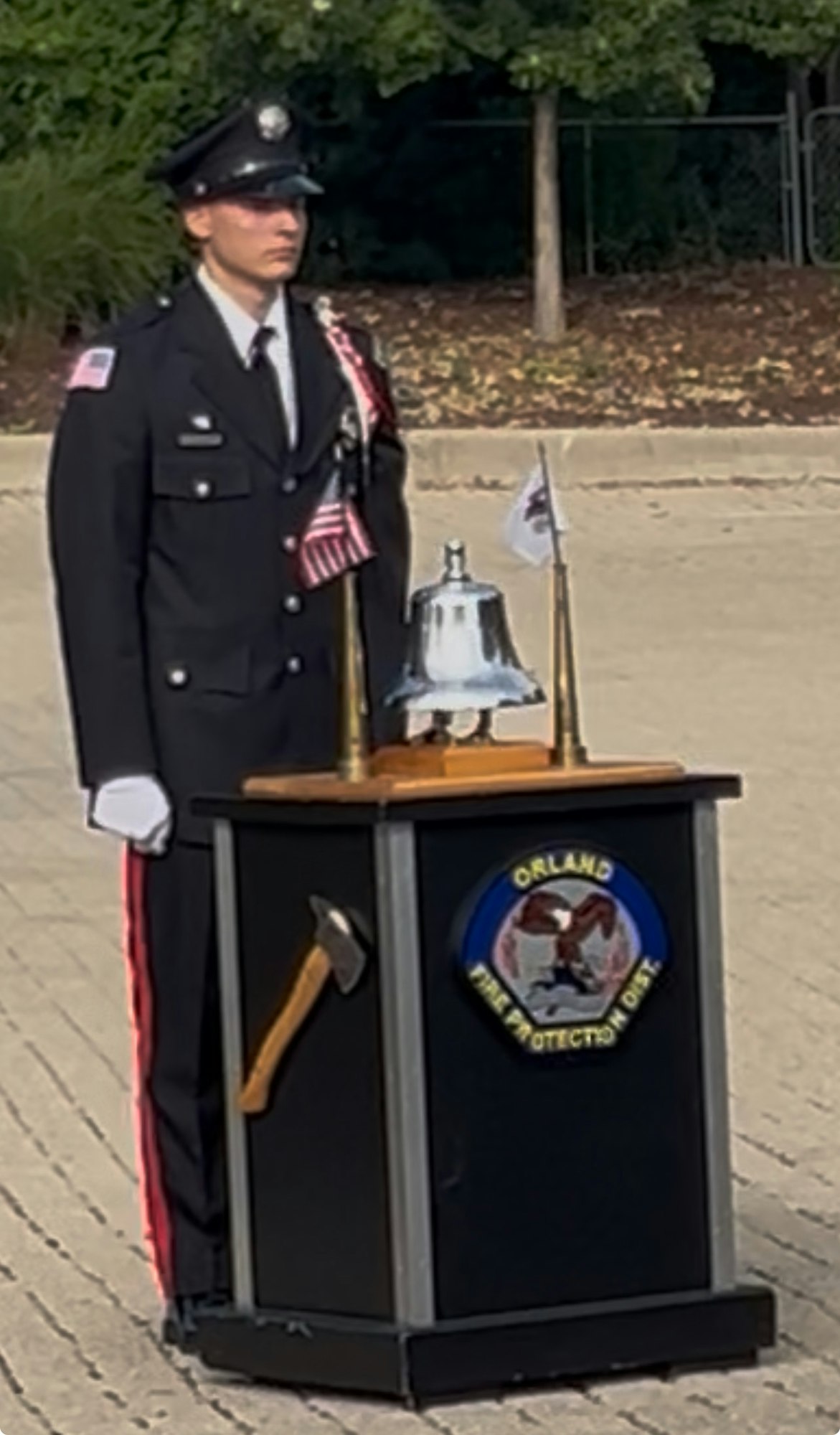 A uniformed officer stands beside a podium with an axe, bell, and flags at an outdoor ceremony.