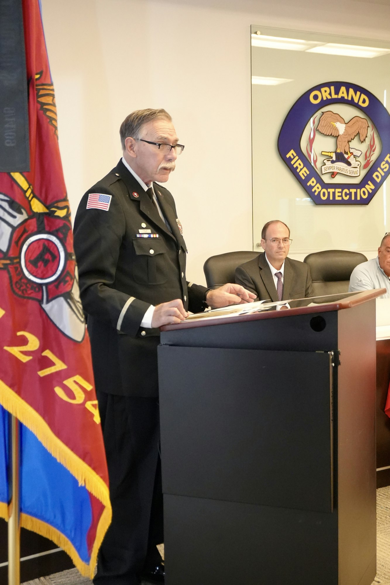 A person in a uniform speaks at a podium with an Orland Fire Protection District emblem and flag visible.