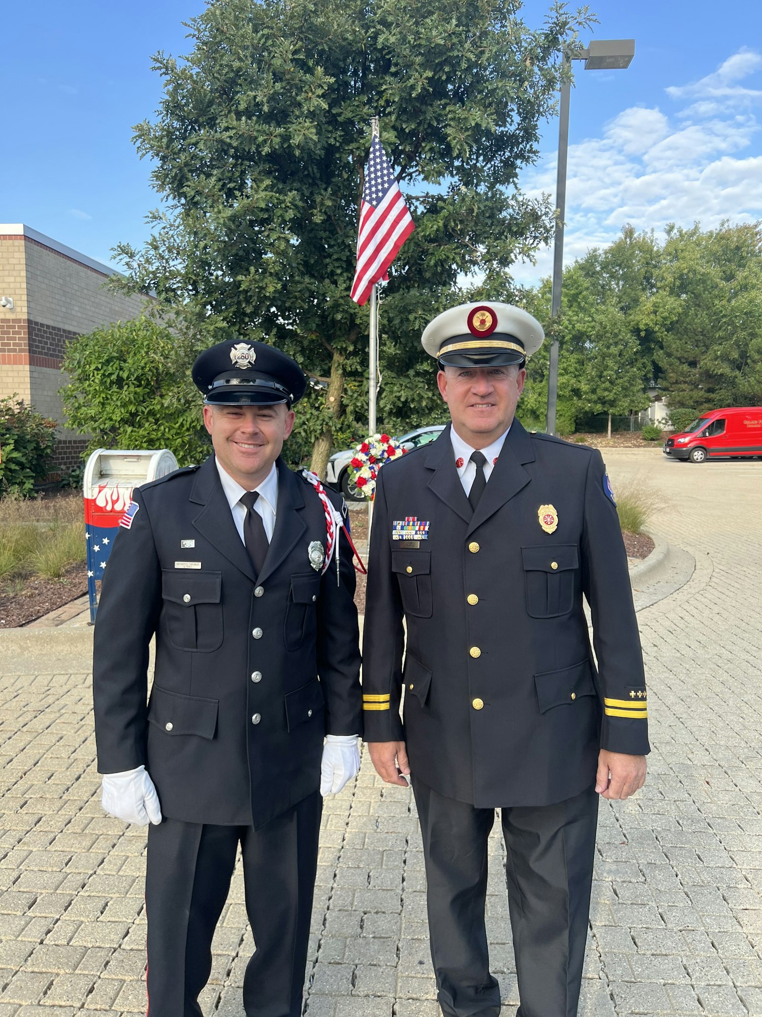 Two people in dress uniforms stand in front of an American flag and a wreath, outdoors on a sunny day.