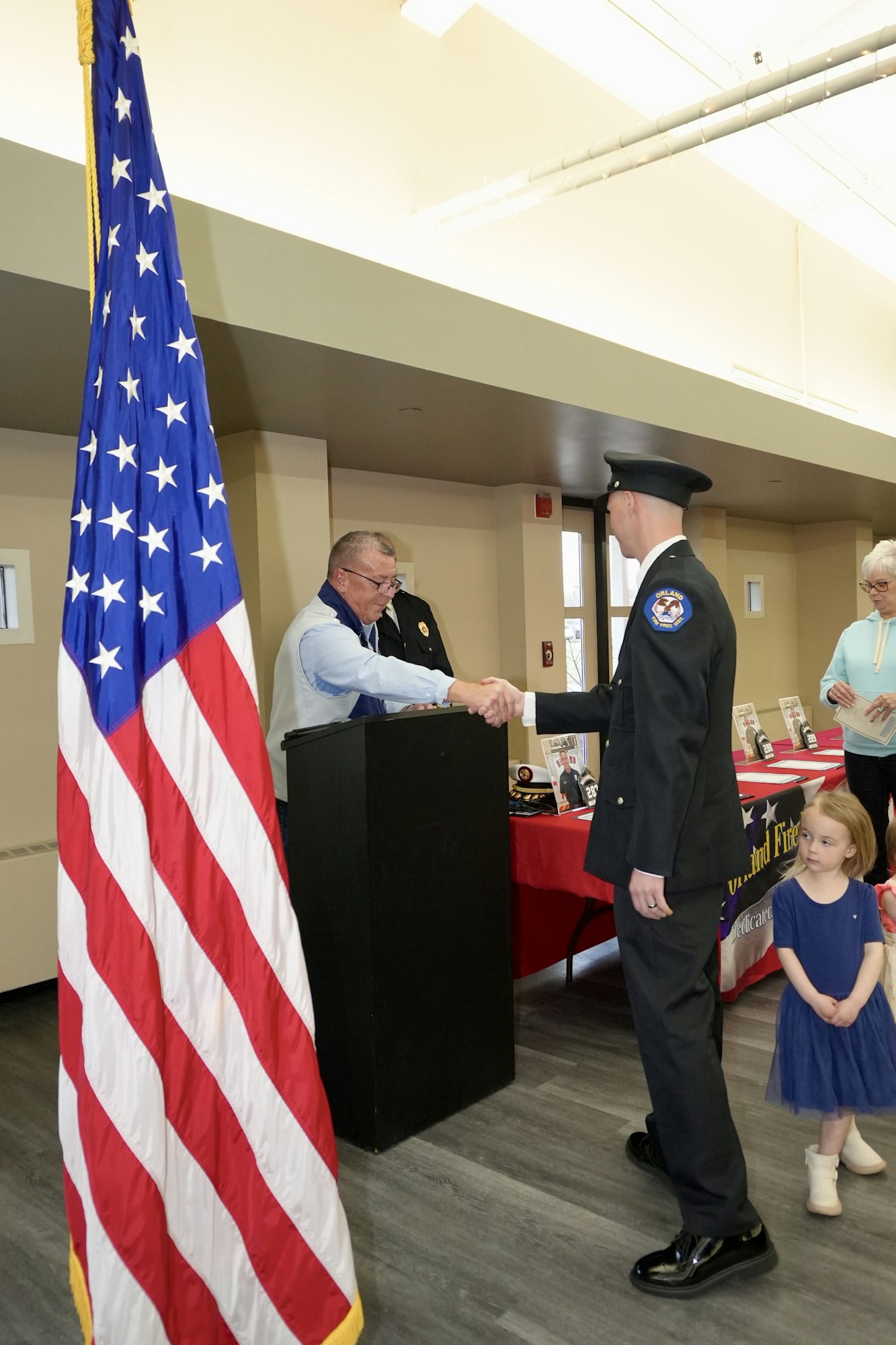 A uniformed man shakes hands with another at a podium beside a flag, with onlookers nearby, including a child.