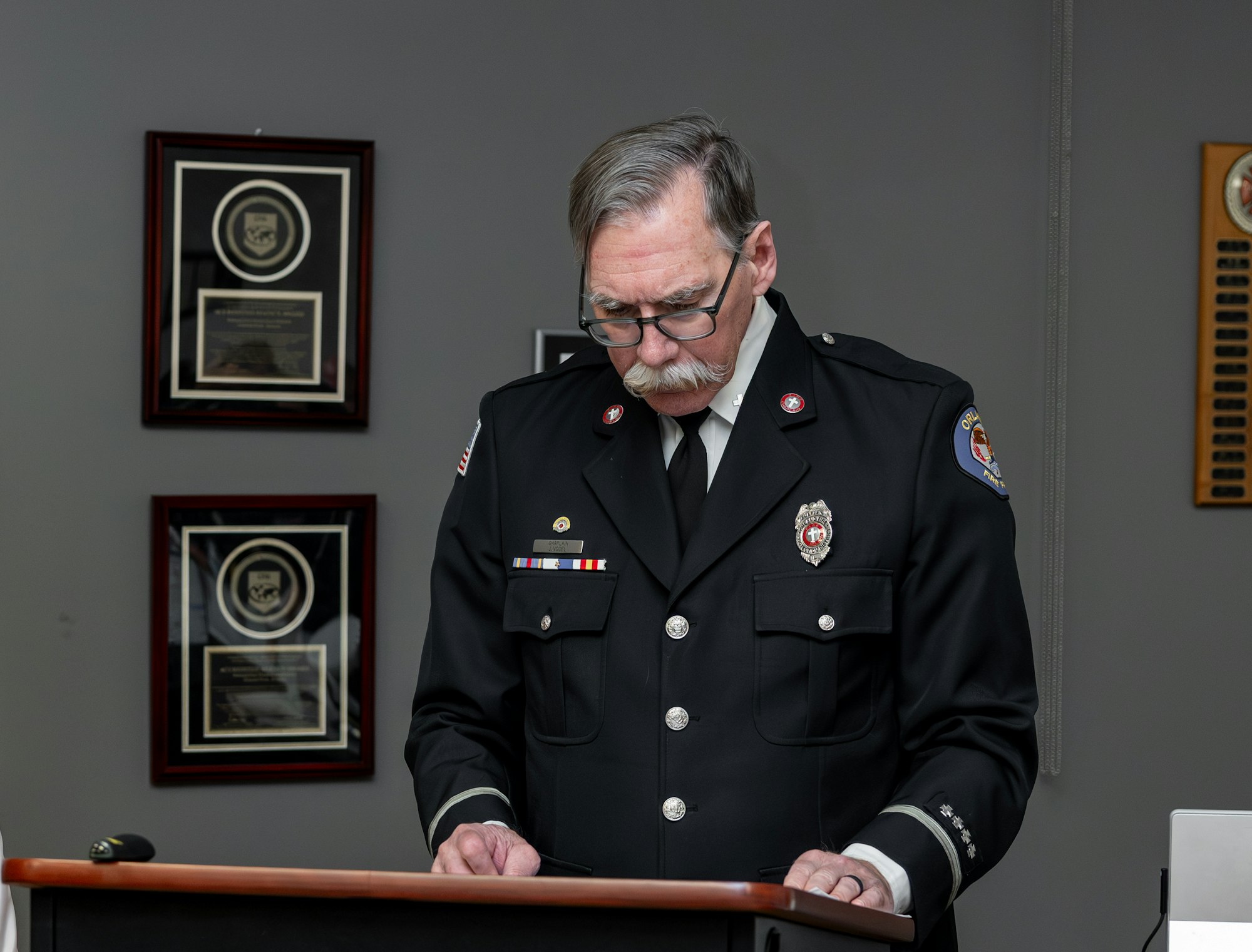 An officer in uniform stands at a podium, looking down, with framed awards displayed on the wall behind him.