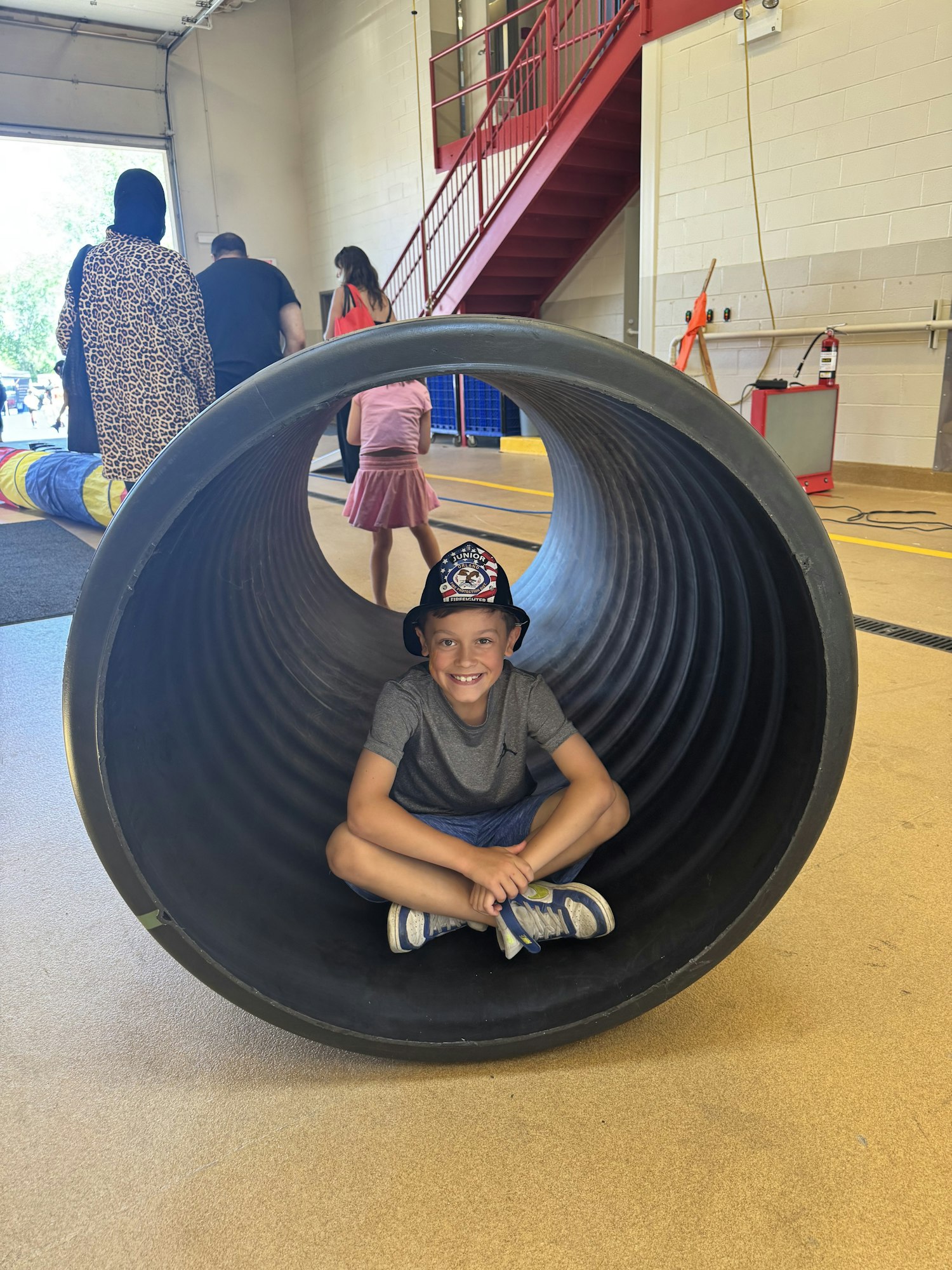 A child wearing a hat and sneakers is sitting inside a large black tube, smiling. People are walking in the background.