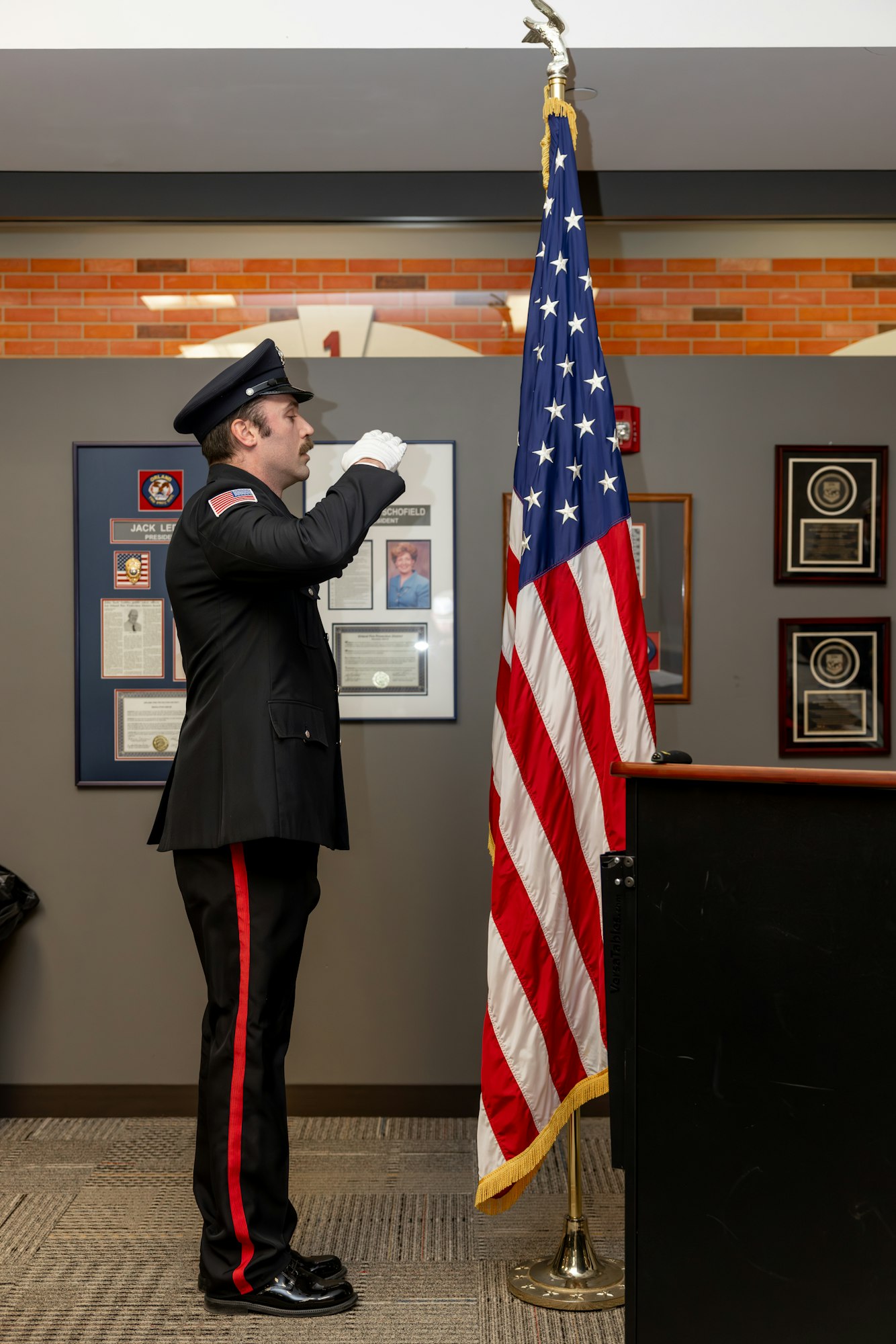 A uniformed officer salutes the American flag in a setting with plaques and a podium.