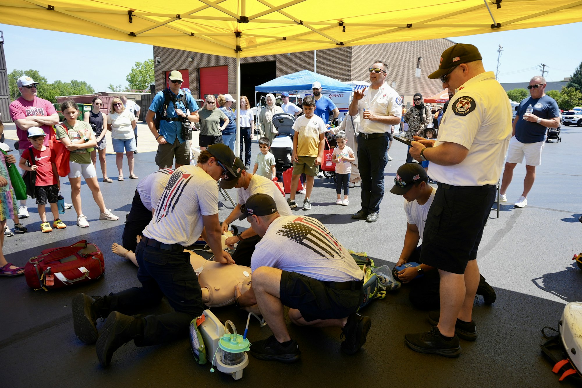 A group is demonstrating CPR on a dummy under a yellow tent, watched by an attentive crowd.