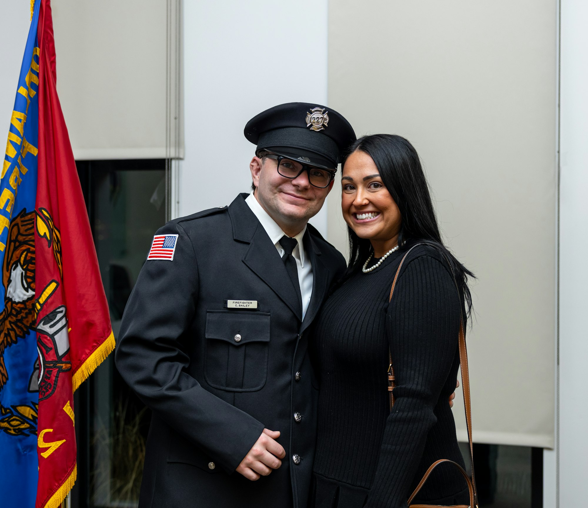 A man in a police uniform poses with a woman, both smiling, with a flag in the background.