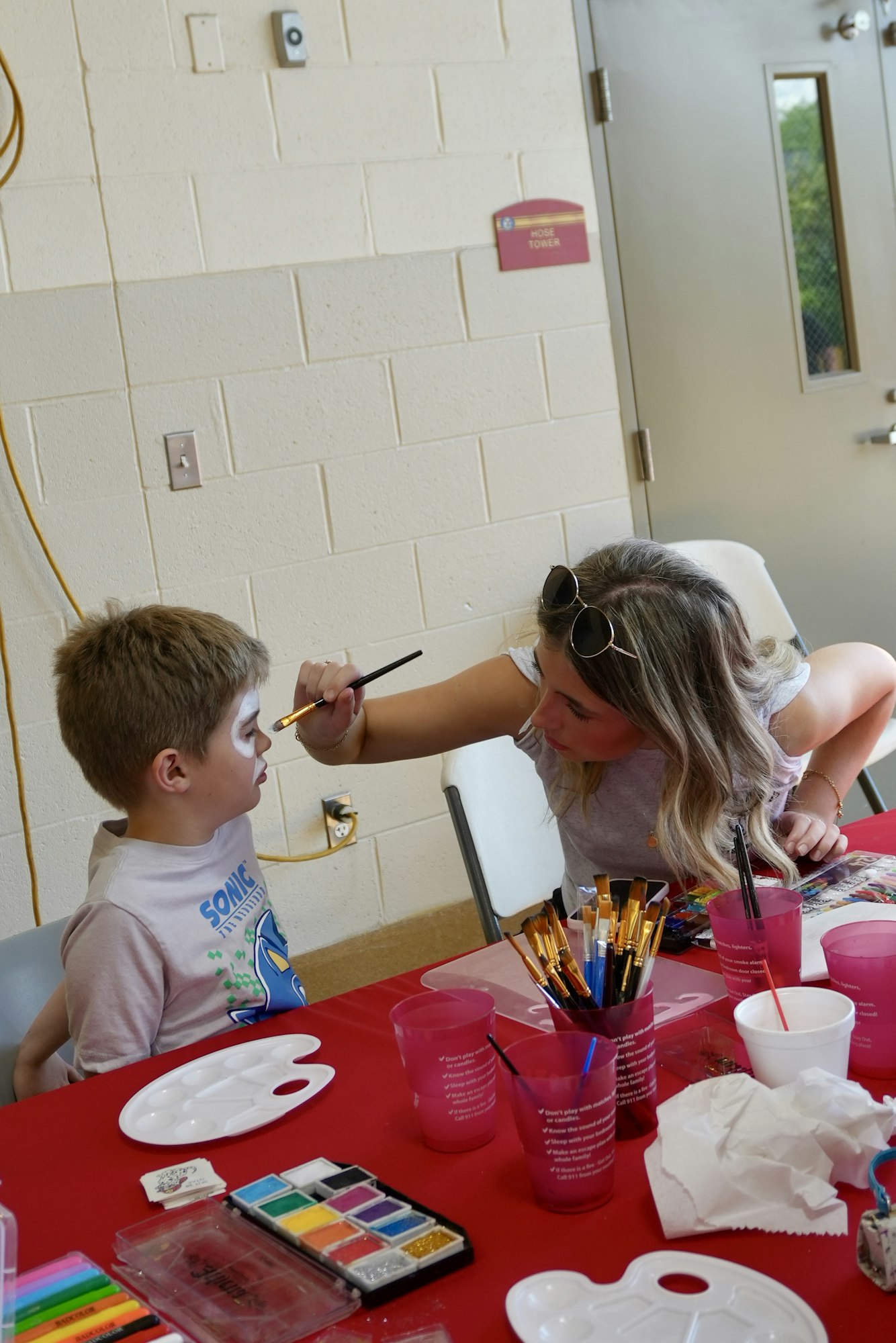 A woman is face painting a young boy at a table with art supplies.
