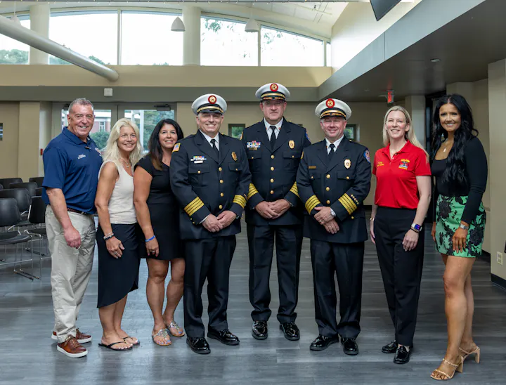 The image shows a group of people, including uniformed officials and civilians, posing together in a bright indoor setting.