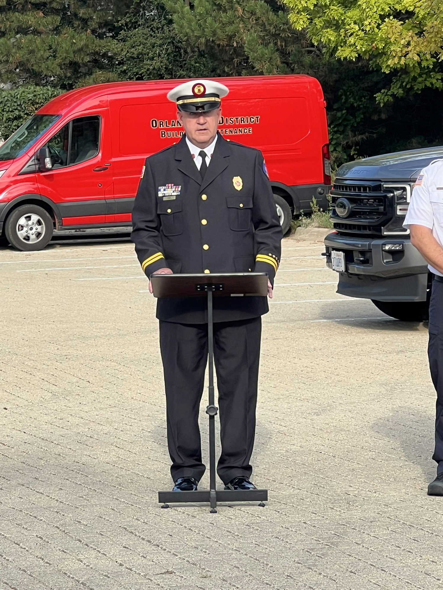 A uniformed person stands at a podium outdoors. A red van and another person are in the background.