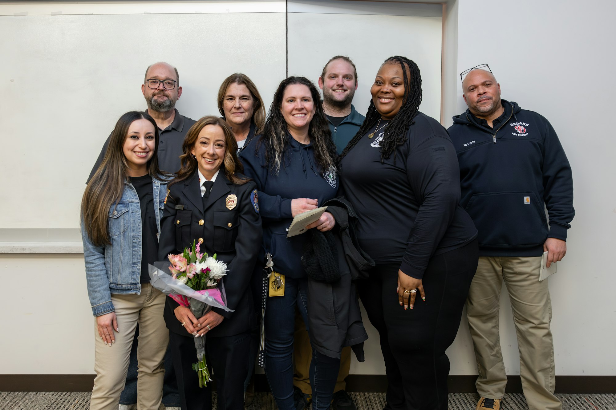 The image shows a group of people, including a smiling woman in a police uniform holding flowers, posing together in a celebratory setting.