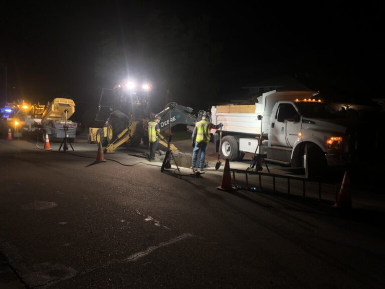 Night roadwork scene with workers, vehicles, and equipment illuminated by bright lights, surrounded by traffic cones.
